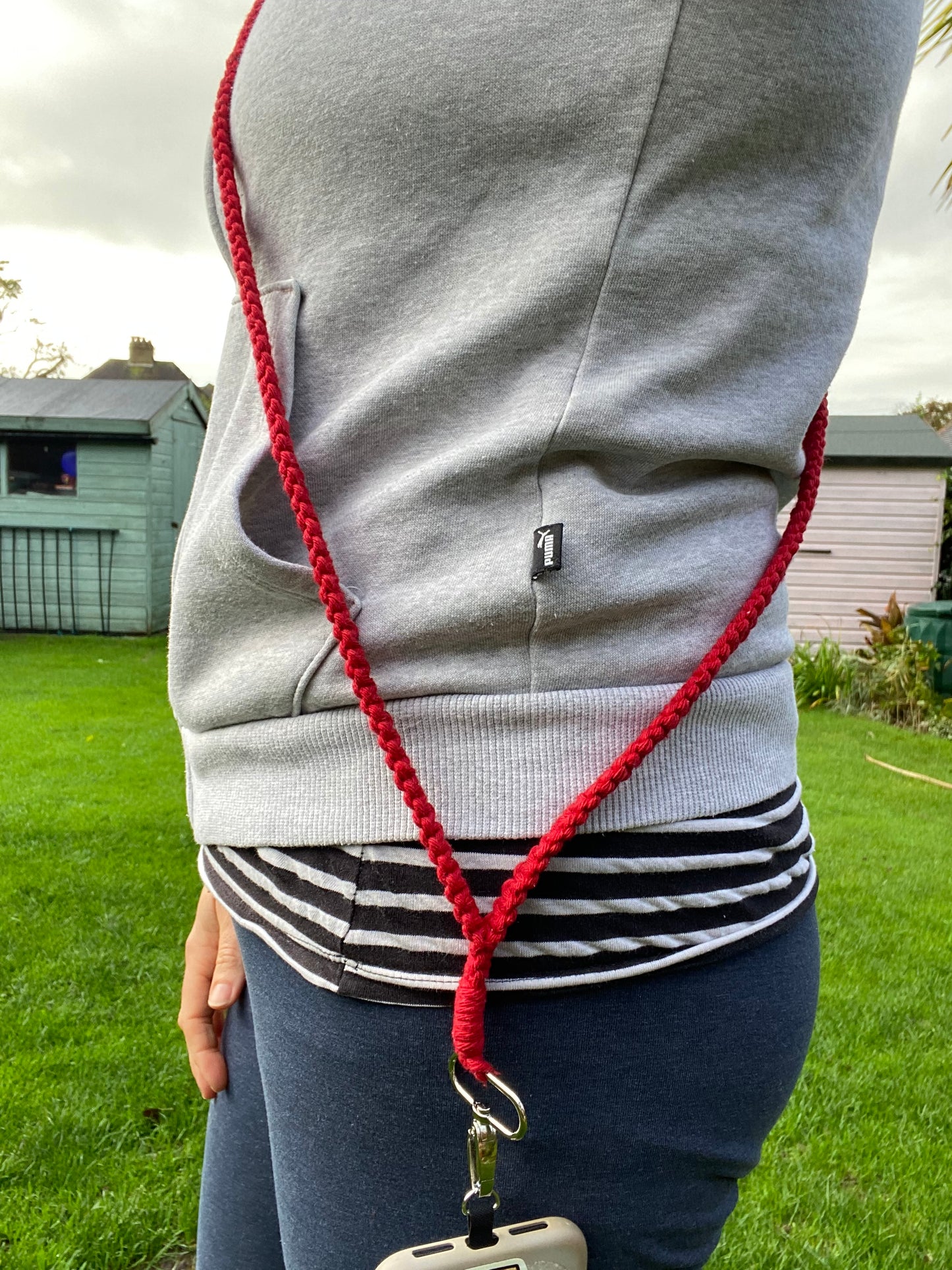 A person is wearing a grey hoodie and dark pants, with a red lanyard hanging from their neck. The lanyard, a Recycled cotton crossbody phone strap by Macra-Made-With-Love, attaches to a phone clipped to their right side. They are standing outdoors on a lawn with a shed and a fence in the background.