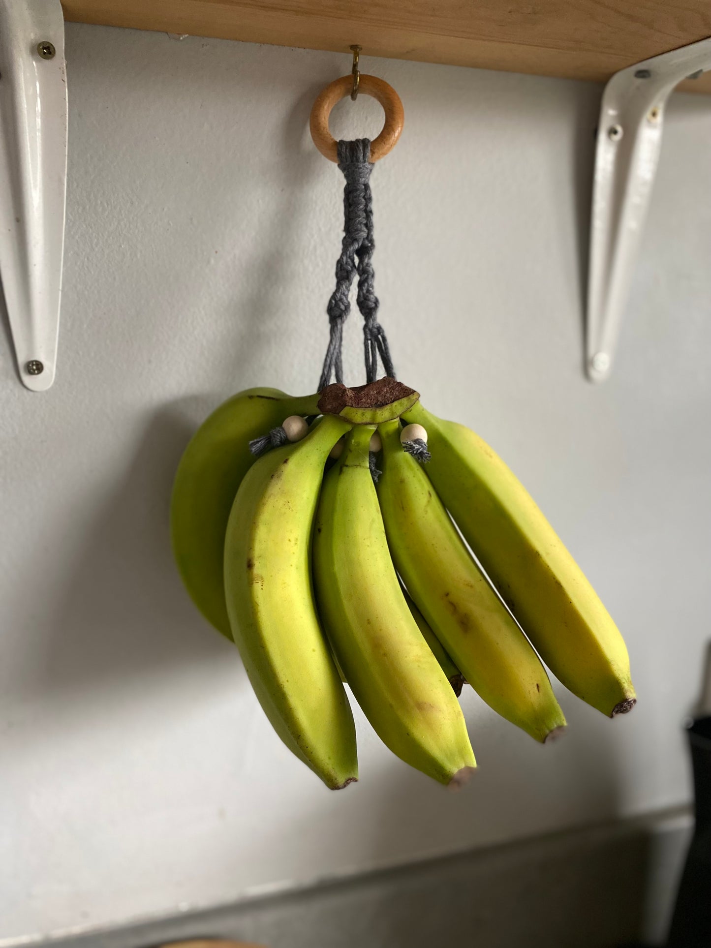 A bunch of yellow bananas hangs from a hand-crafted, macrame-style **Banana Hanger** by **Macra-Made-With-Love** attached to the underside of a wooden shelf. This eco-friendly home decor piece is decorated with beads and a wooden ring. The background features a white wall, and part of the countertop is visible at the bottom.