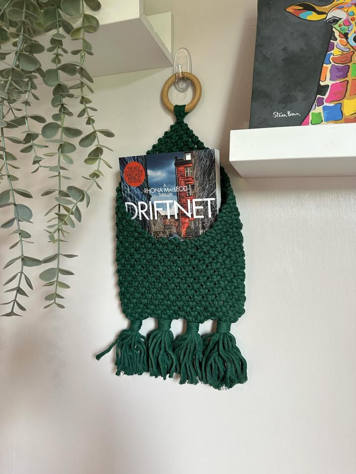 A green crocheted wall pouch made of recycled cotton decor with tassels hangs on a hook beside a leafy plant. The handmade Macra-Made-With-Love Bedside Storage Pouch Book Sleeve holds a book titled "Drift Net" by Rhona MacLeod. Above the pouch is a white shelf with a colorful giraffe painting on display.