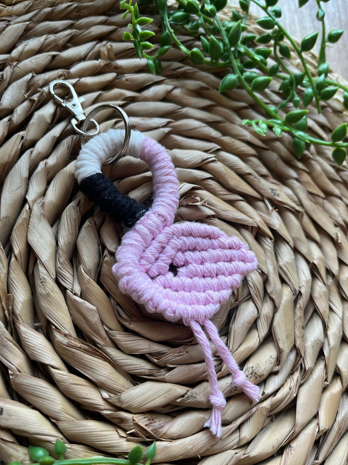 Handmade macrame keychain in the shape of a flamingo, made using light pink recycled cotton, on a metal split ring with lobster clasp. The keyring is displayed on a round wicker surface next to some green plant vines.