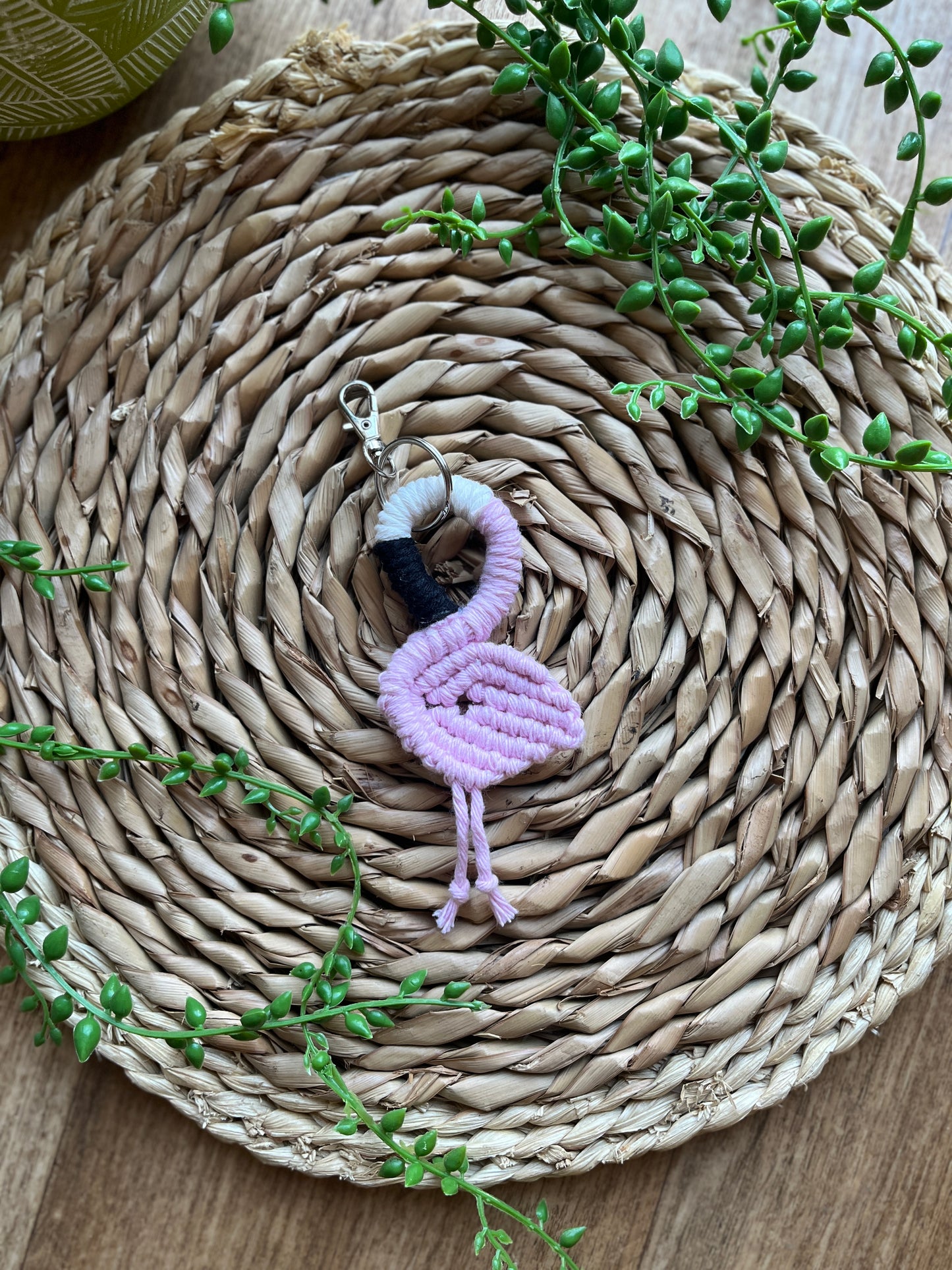 Handmade macrame keychain in the shape of a flamingo, made using light pink recycled cotton, on a metal split ring with lobster clasp. The keyring is displayed on a round wicker surface next to some green plant vines.