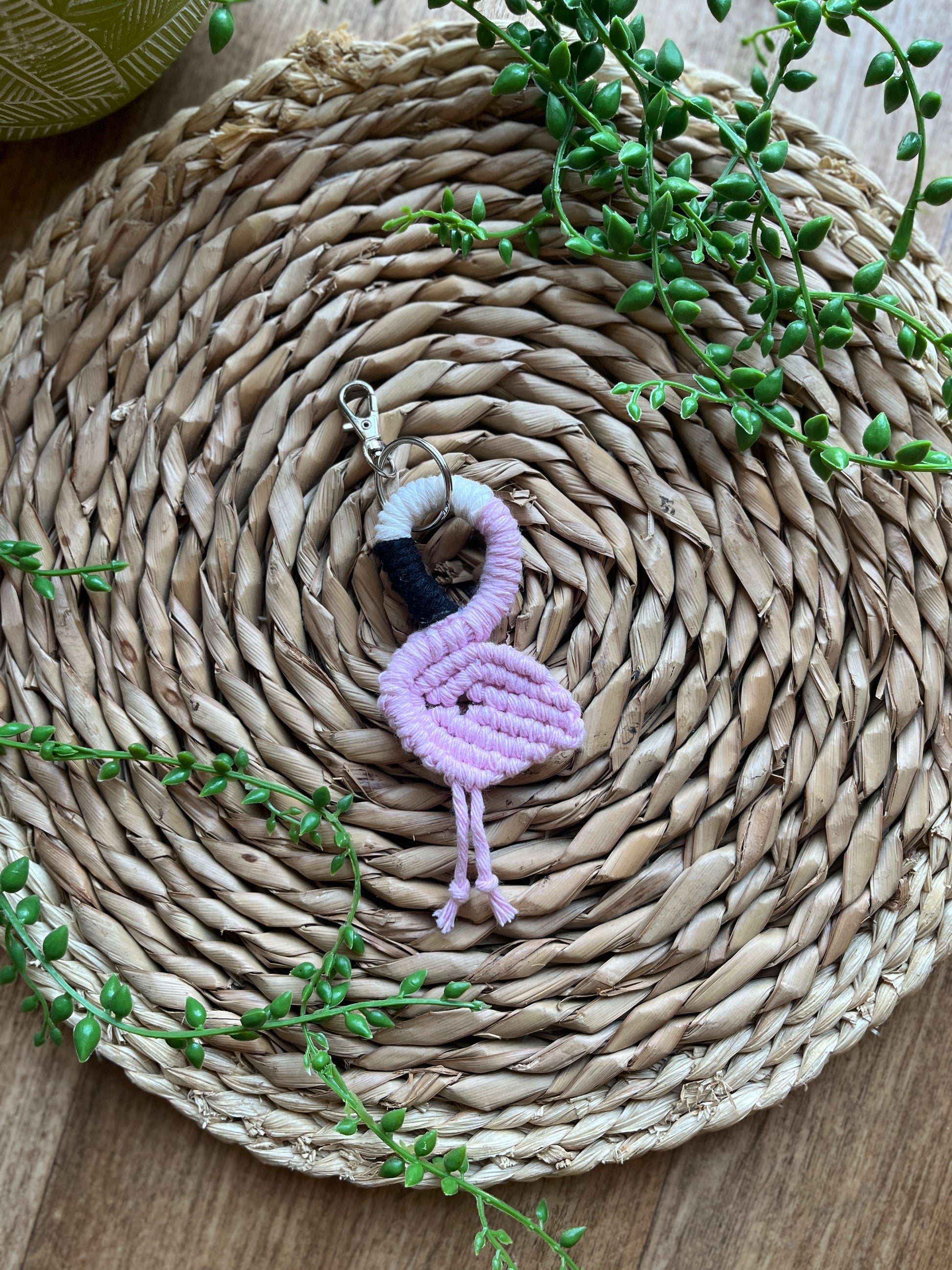 Handmade macrame keychain in the shape of a flamingo, made using light pink recycled cotton, on a metal split ring with lobster clasp. The keyring is displayed on a round wicker surface next to some green plant vines.