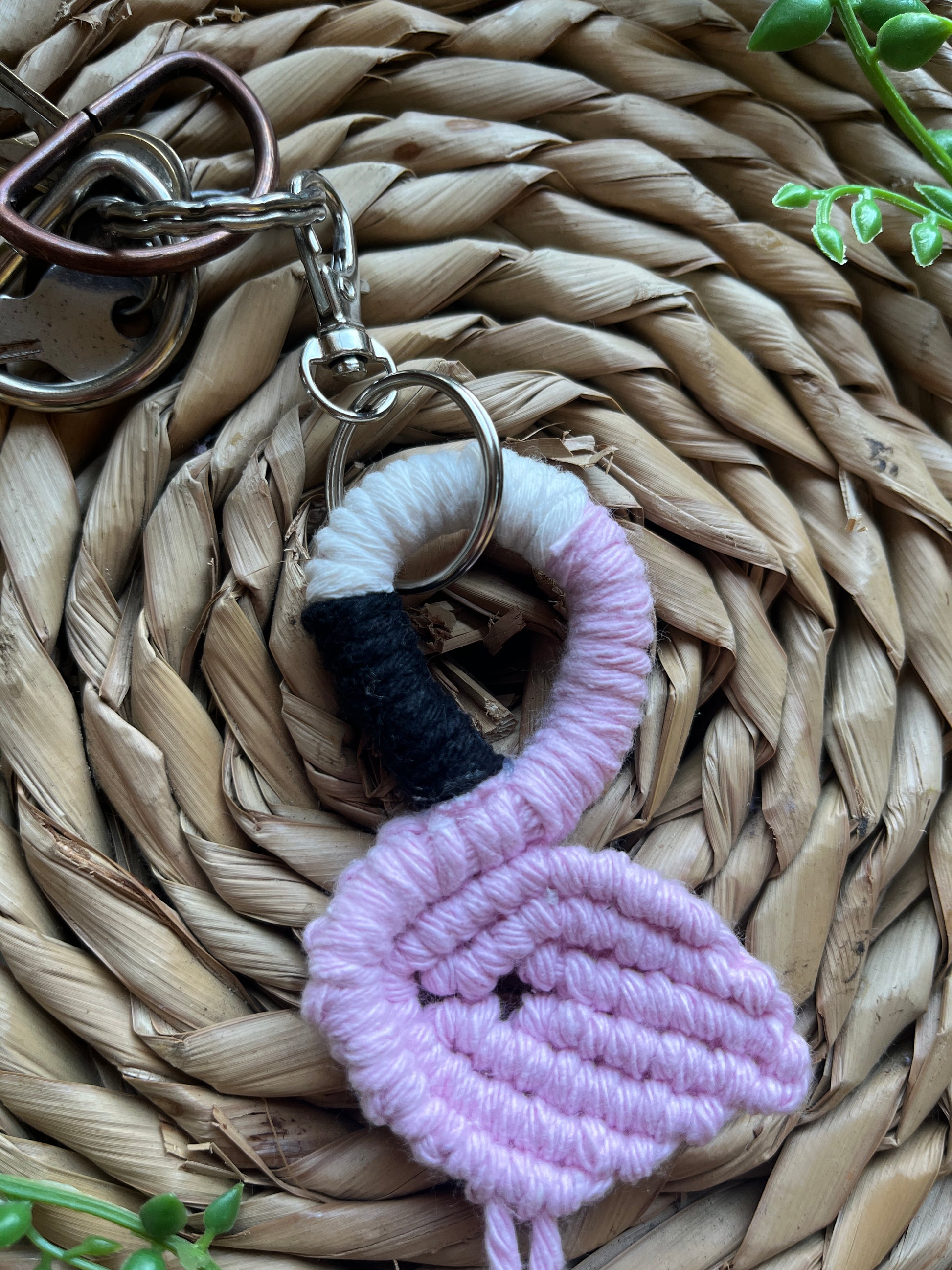 Handmade macrame keychain in the shape of a flamingo, made using light pink recycled cotton, on a metal split ring with lobster clasp. The keyring is displayed on a round wicker surface next to some green plant vines.