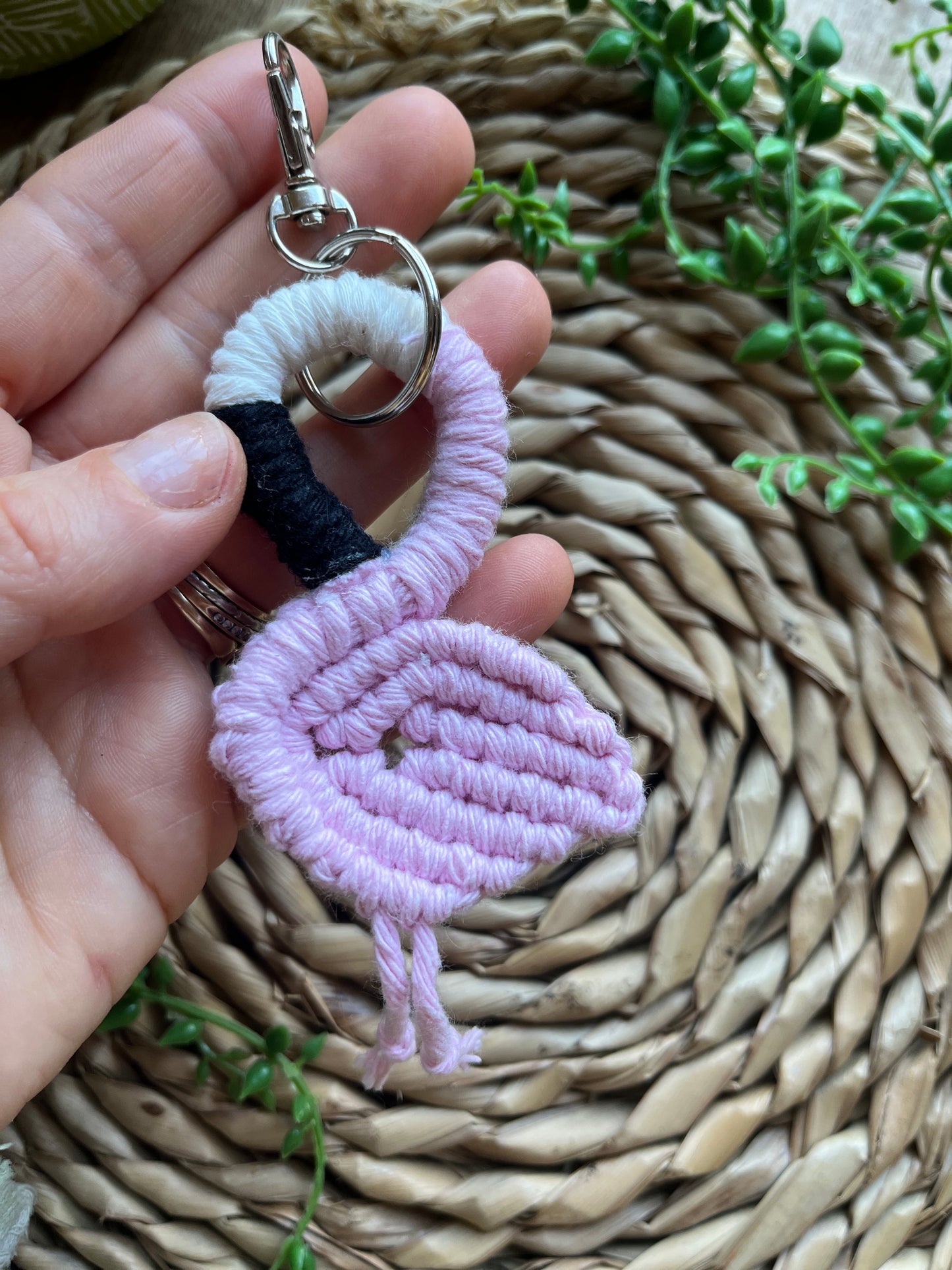 A hand holding a handmade macrame keychain in the shape of a flamingo, made using light pink recycled cotton, on a metal split ring with lobster clasp. The keyring is displayed over a round wicker surface next to some green plant vines.