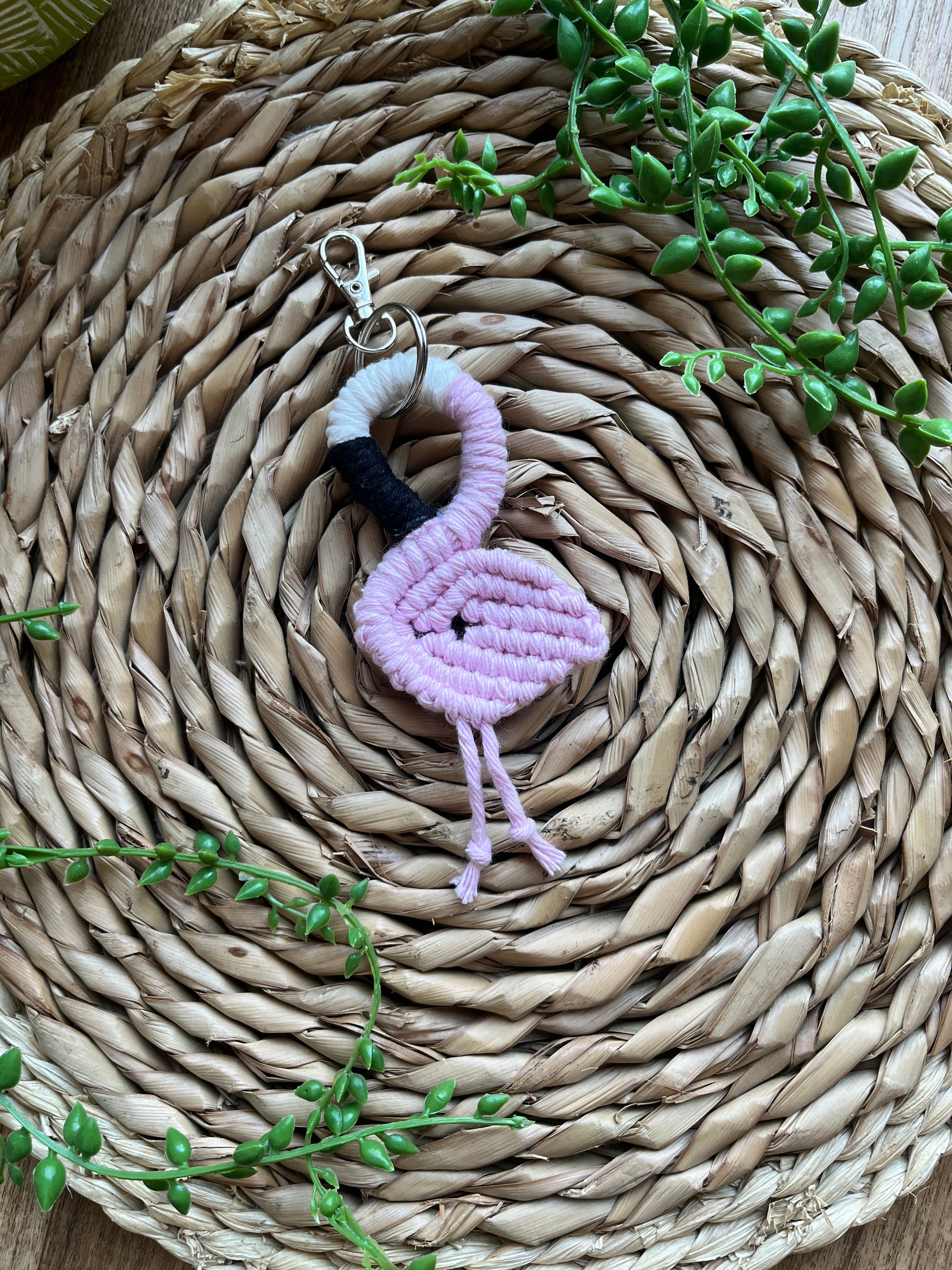 Handmade macrame keychain in the shape of a flamingo, made using light pink recycled cotton, on a metal split ring with lobster clasp. The keyring is displayed on a round wicker surface next to some green plant vines.