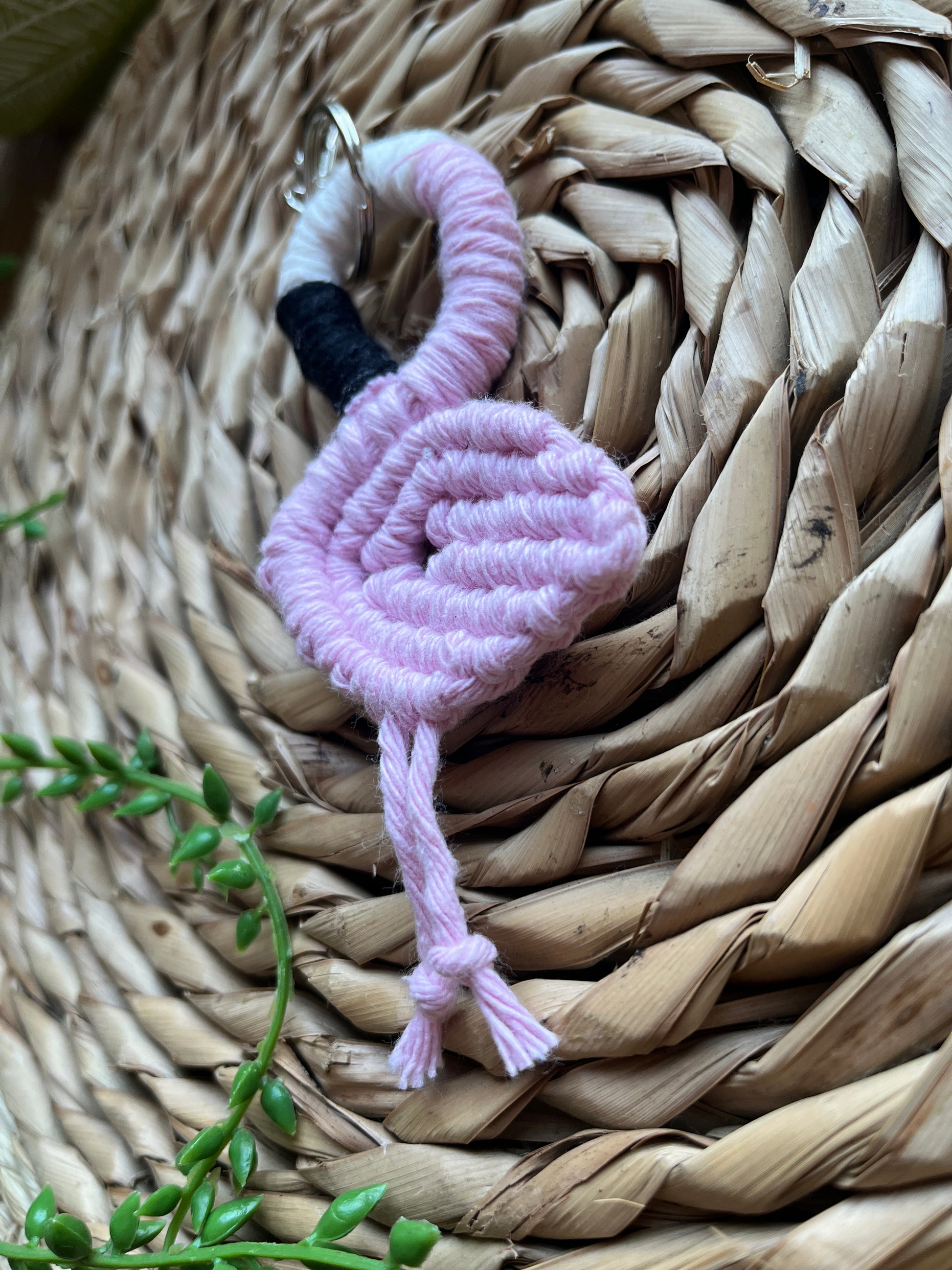 Handmade macrame keychain in the shape of a flamingo, made using light pink recycled cotton, on a metal split ring with lobster clasp. The keyring is displayed on a round wicker surface next to some green plant vines.