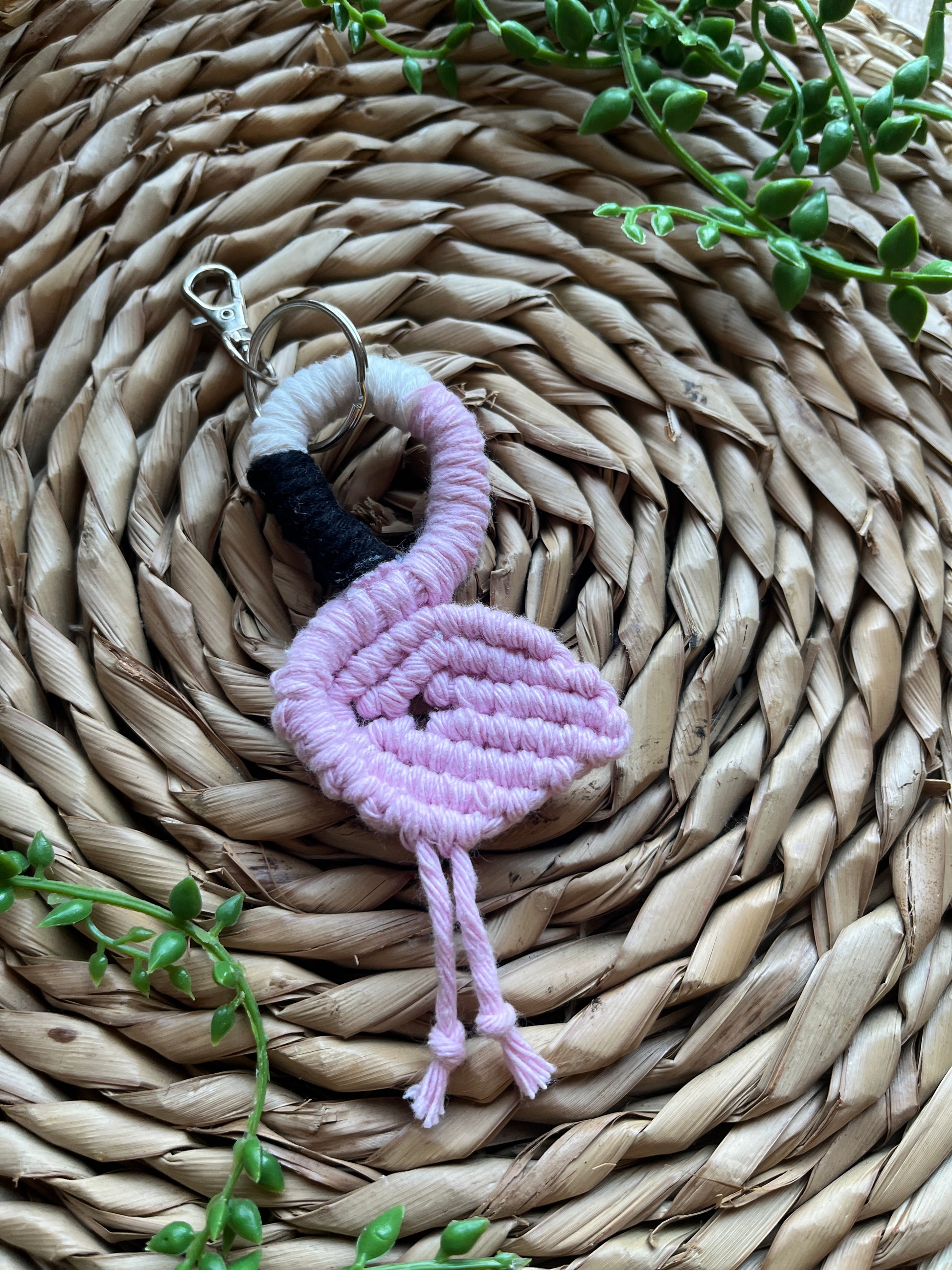 Handmade macrame keychain in the shape of a flamingo, made using light pink recycled cotton, on a metal split ring with lobster clasp. The keyring is displayed on a round wicker surface next to some green plant vines.
