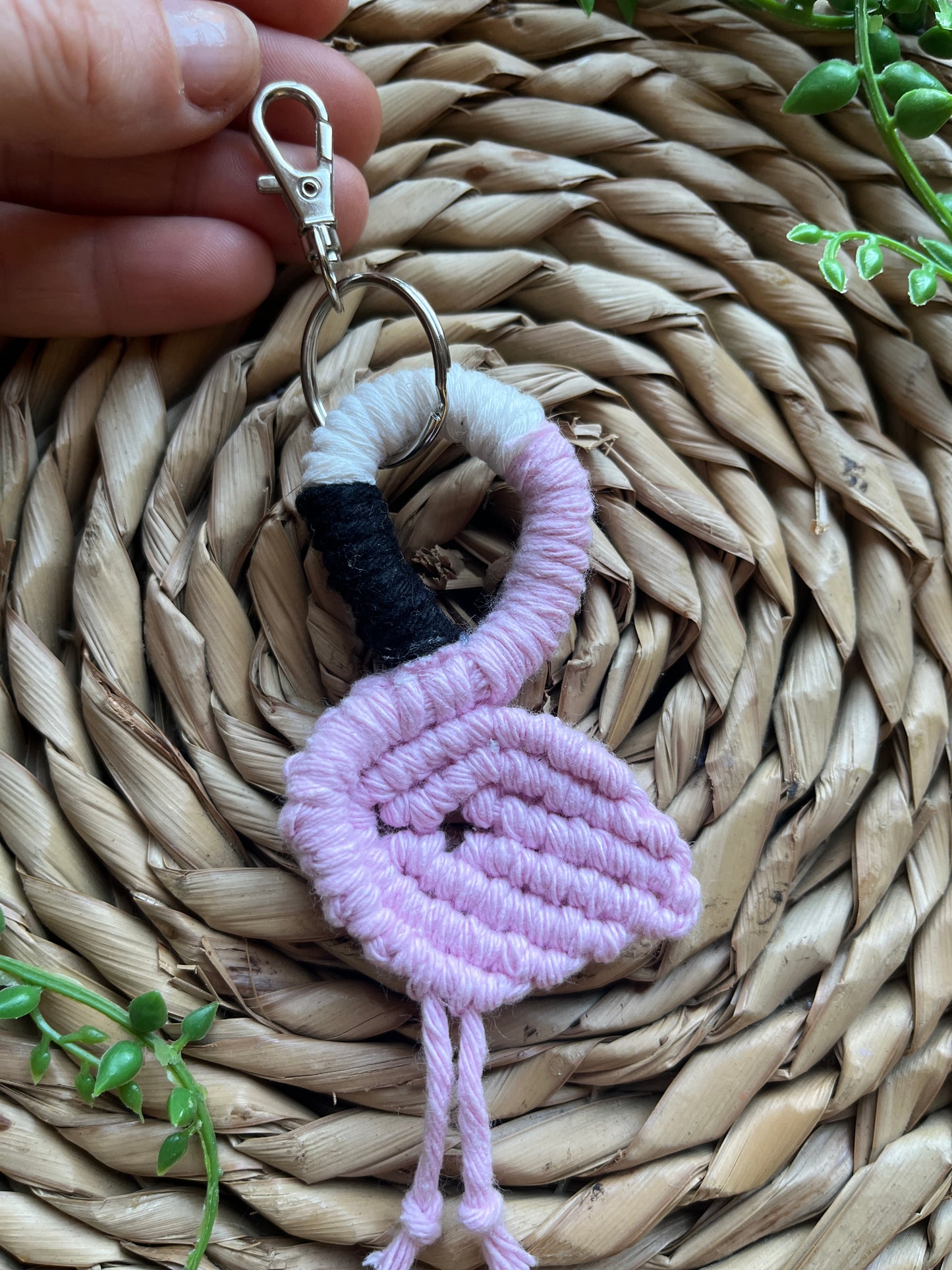Handmade macrame keychain in the shape of a flamingo, made using light pink recycled cotton, on a metal split ring with lobster clasp. The keyring is displayed on a round wicker surface next to some green plant vines.