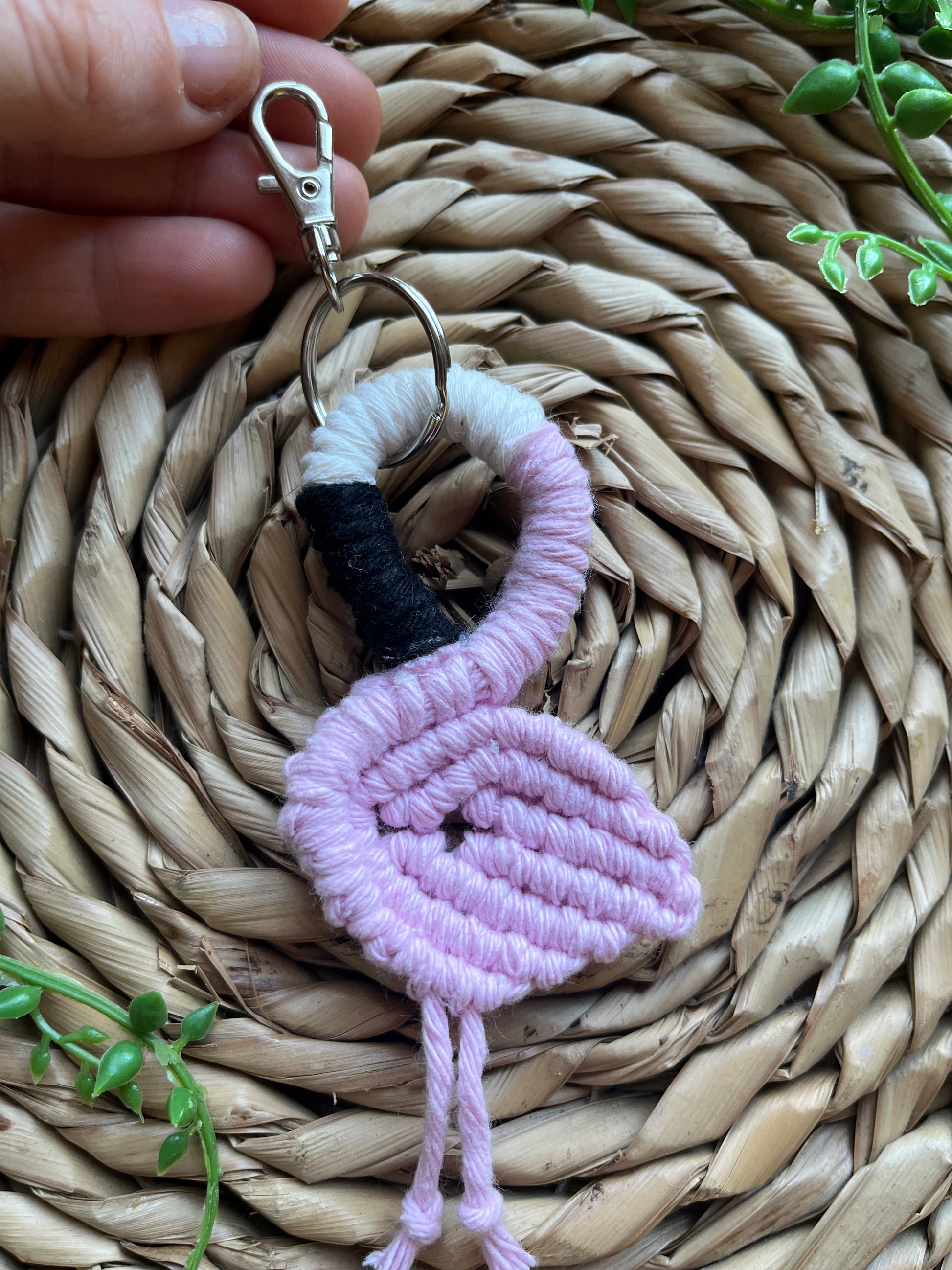 Handmade macrame keychain in the shape of a flamingo, made using light pink recycled cotton, on a metal split ring with lobster clasp. The keyring is displayed on a round wicker surface next to some green plant vines.