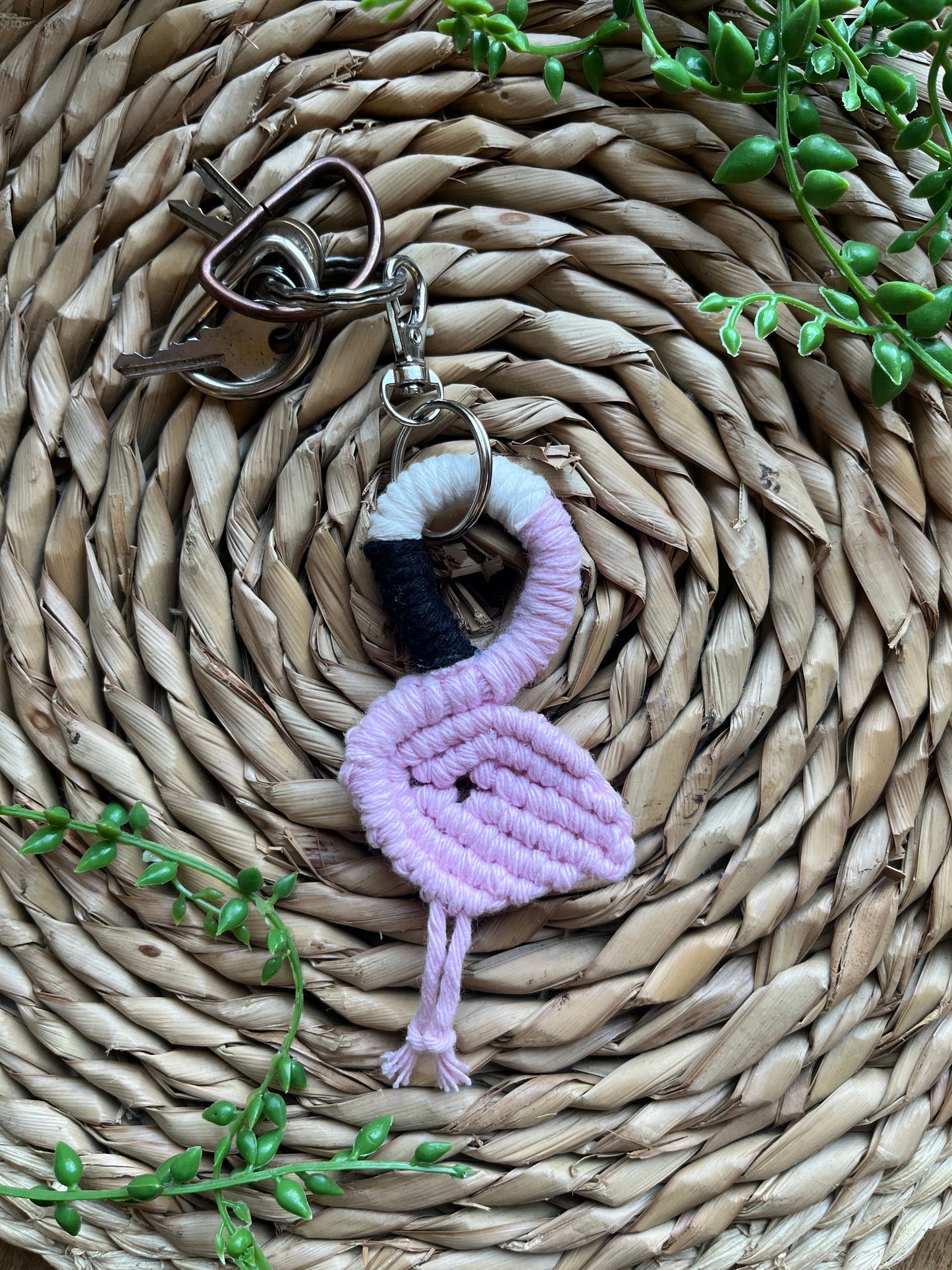 Handmade macrame keychain in the shape of a flamingo, made using light pink recycled cotton, on a metal split ring with lobster clasp. The keyring is displayed on a round wicker surface next to some green plant vines.