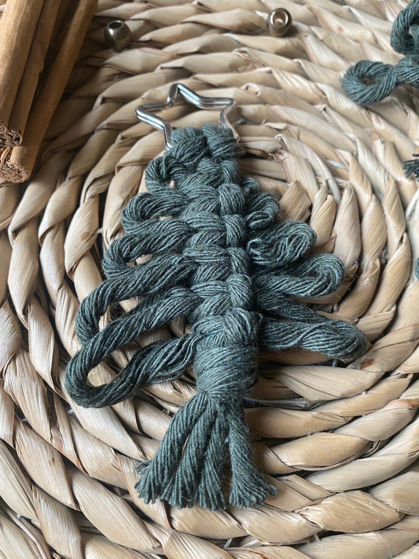 A small, green macrame keychain in the shape of a leaf lies on a woven straw placemat. This eco-friendly holiday decor piece, made from recycled cotton yarns and known as the Knotted Yarn Hanging Christmas Tree Decoration from Macra-Made-With-Love, adds charm to any setting. To the left, two cinnamon sticks are partially visible and threads can be seen in the background.