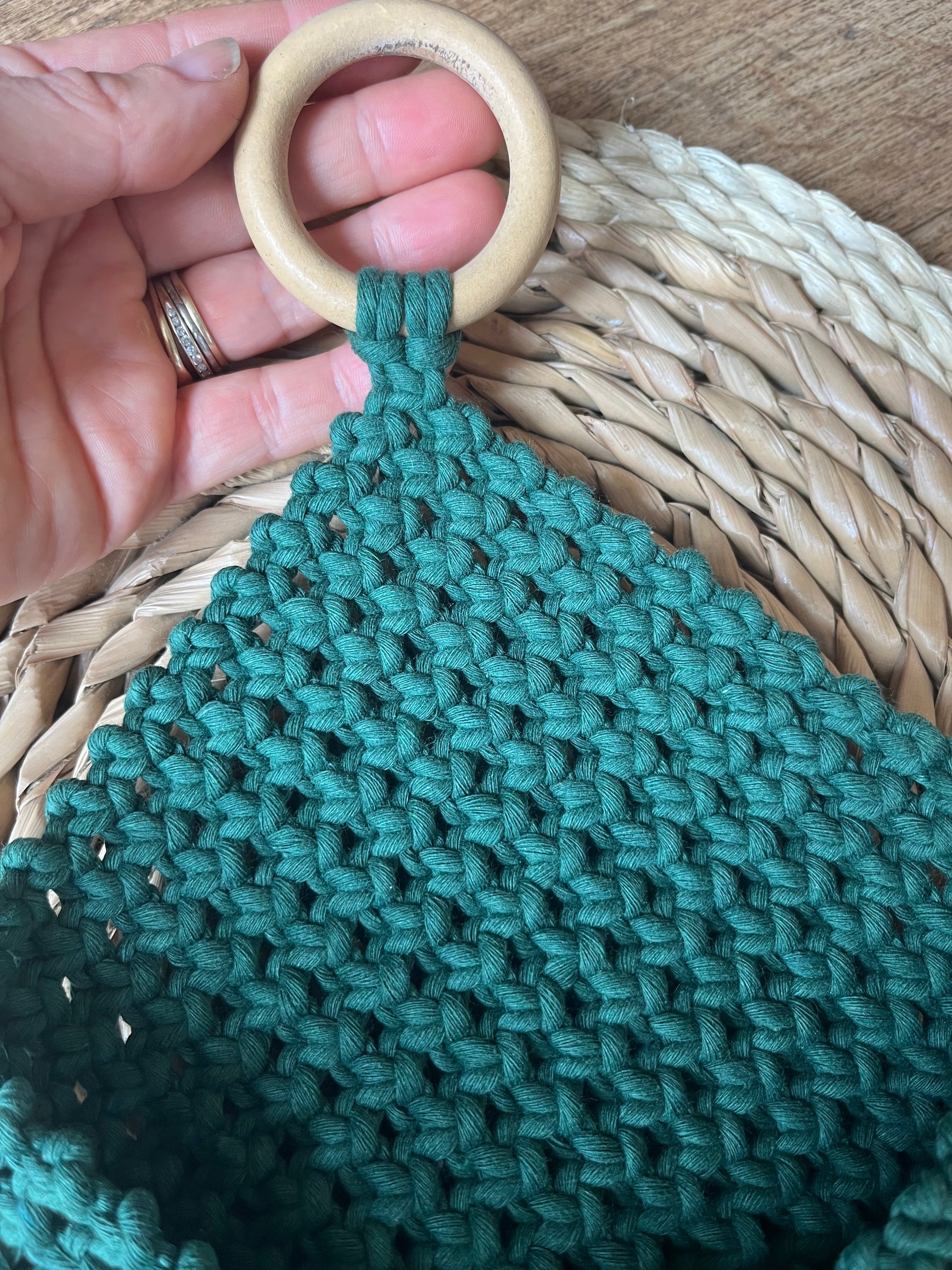 A hand holds a wooden ring attached to a green, woven fabric. The fabric has a textured, crochet-like pattern. The background includes a woven straw mat on a wooden surface. The hand is adorned with a silver ring, showcasing the Macra-Made-With-Love Bedside Storage Pouch Book Sleeve made from recycled cotton decor.