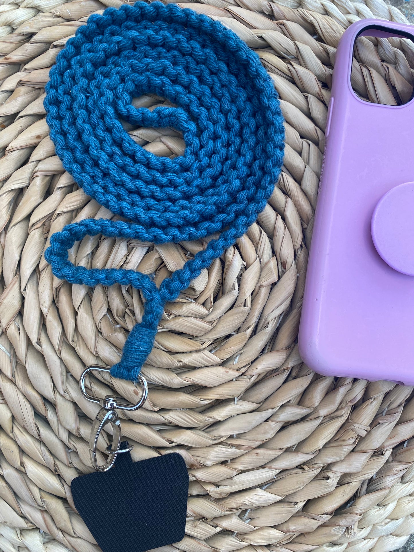 A coiled blue rope lanyard with a metal clip and black attachment sits on a woven circular mat. Next to it is an eco-friendly light purple phone case with a round pop socket and a handmade Recycled cotton crossbody phone strap from Macra-Made-With-Love.