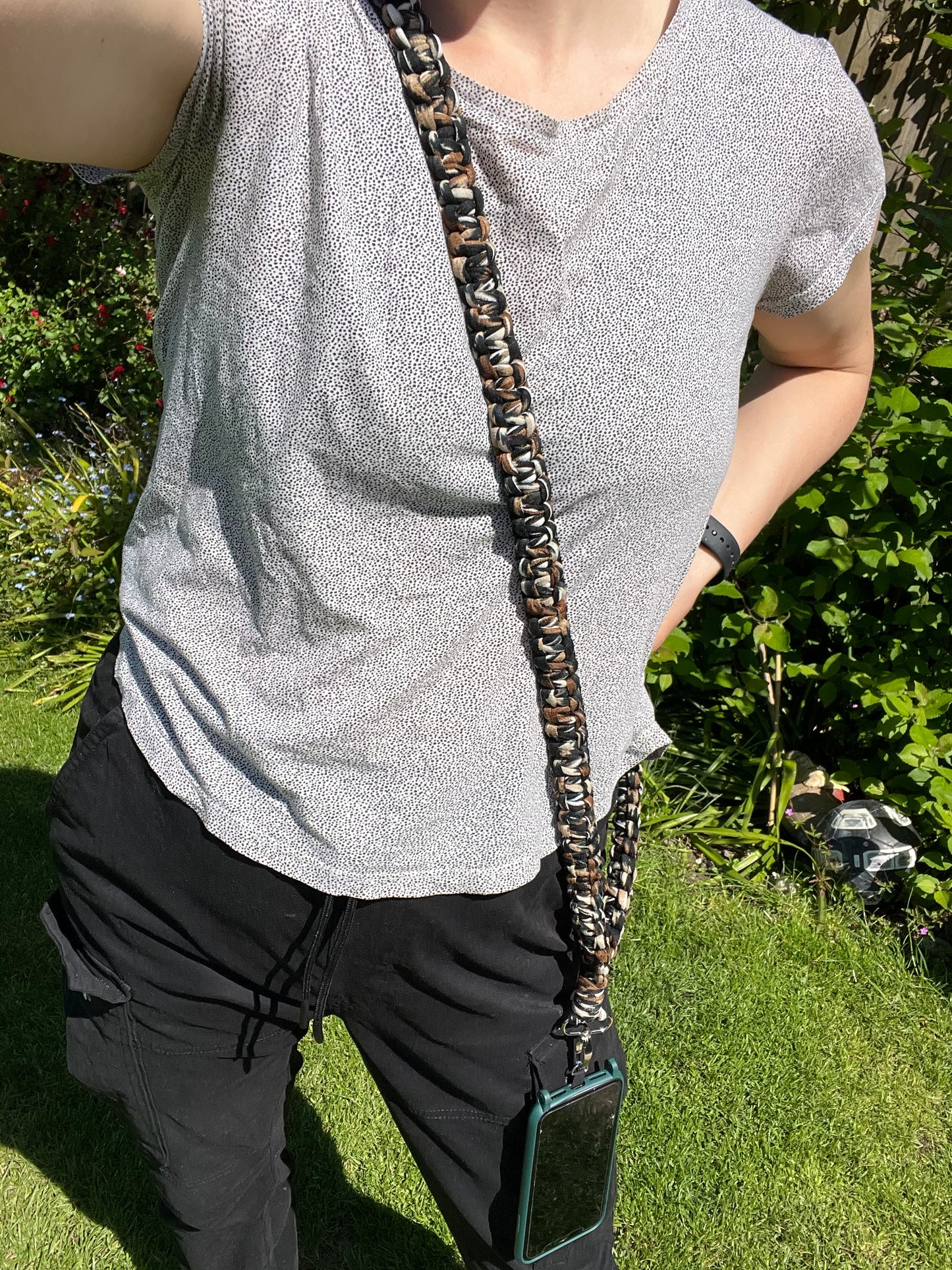 Person wearing a light gray short-sleeve top and black pants, standing on grass. They have a Macra-Made-With-Love Recycled Cotton Crossbody Phone Strap around their neck with a phone attached to it, hanging in front. Part of their arm is visible, with a garden background behind.