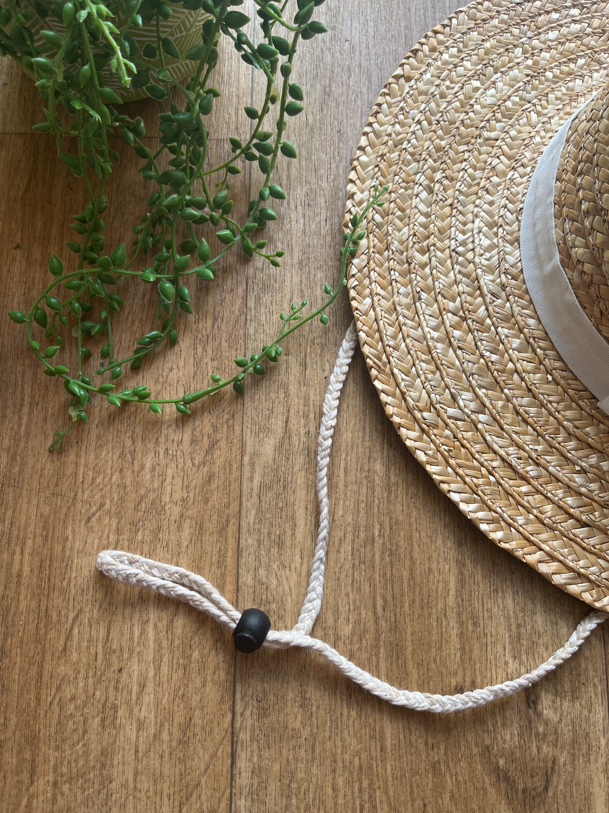 A straw hat with a chin strap lanyard with black toggle, on a wooden surface next to a plant.