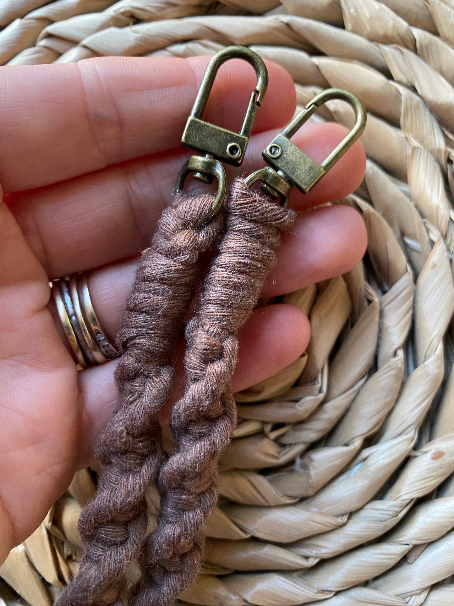 A hand holding the metal clasps on the ends of a long crossbody phone strap, made with a twisted knot pattern in brown yarn.