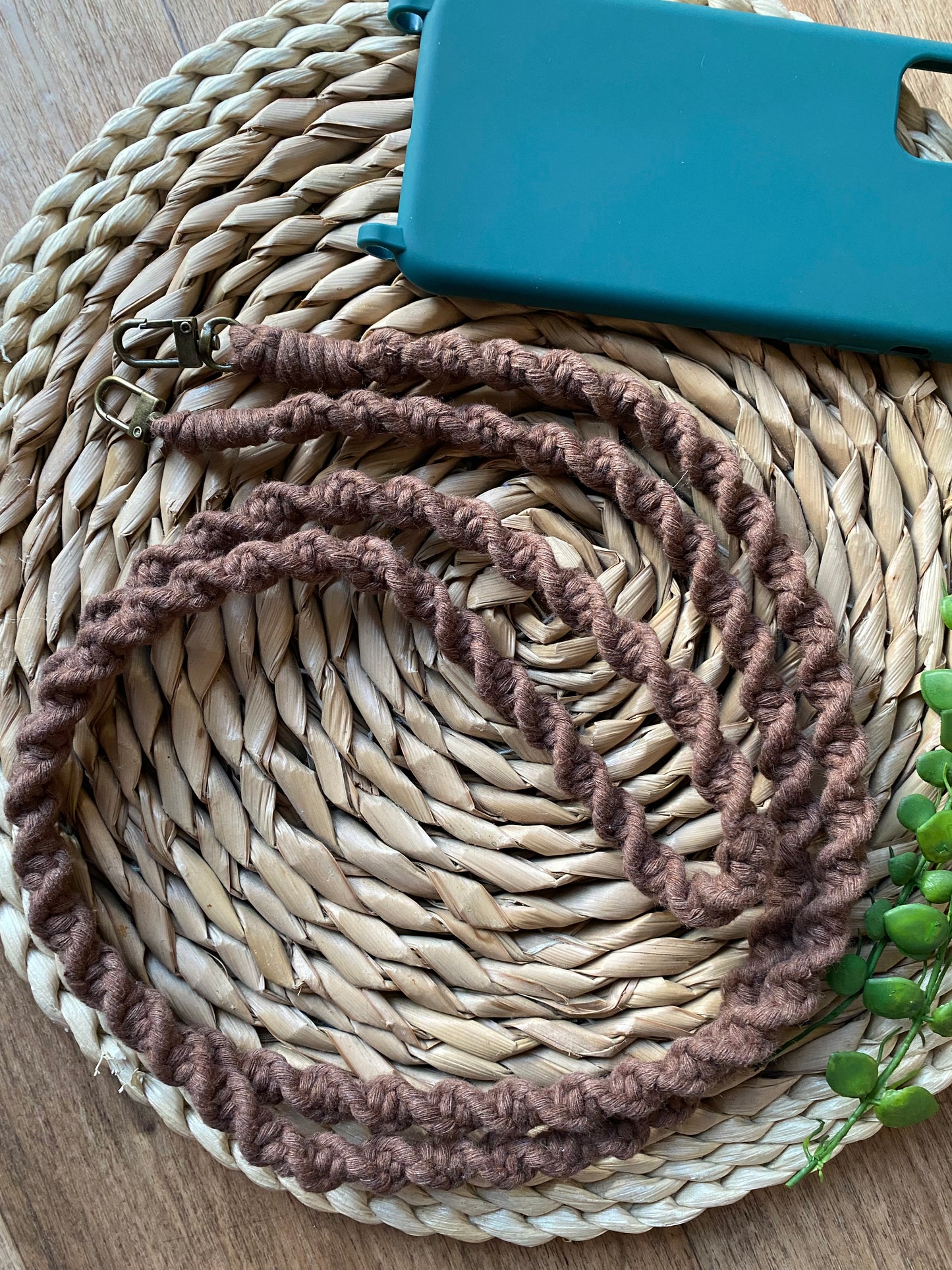 A long macrame crossbody phone strap made with brown yarn and metal clasps, laid neatly on a woven wicker mat next to an empty green phone case.