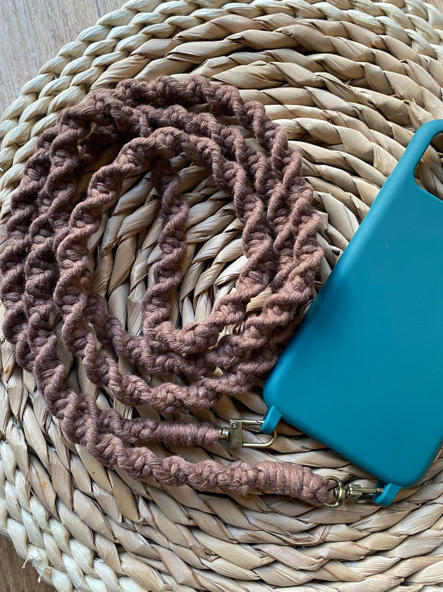 A handmade macrame crossbody phone strap made with brown yarn, neatly coiled on a woven wicker mat, and attached to the bottom of a green phone case.