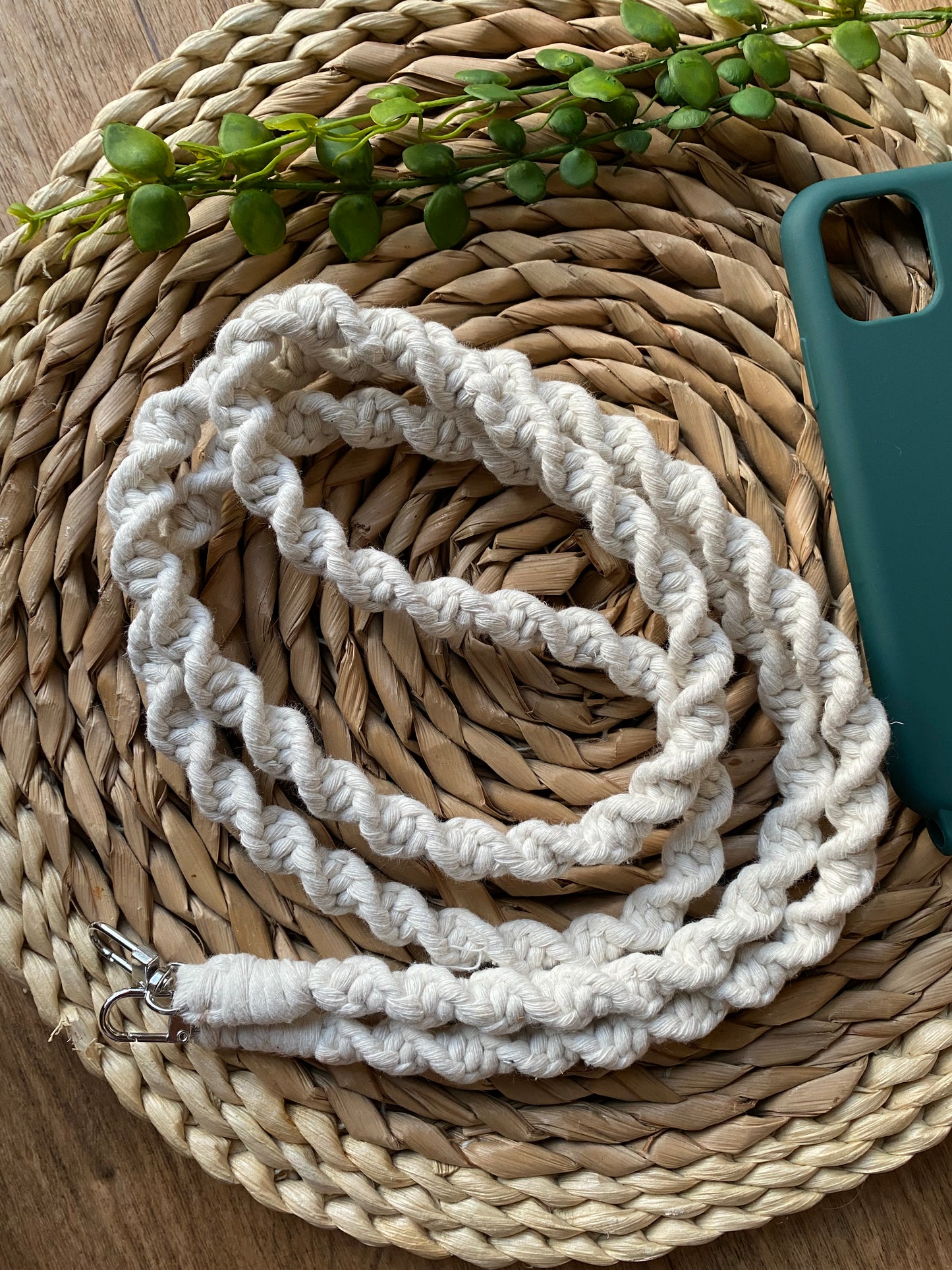 A macrame crossbody phone strap with a twist knot pattern, made with natural yarn and two silver clips on either end, folded in a neat coil on a woven mat next to an empty green phone case.