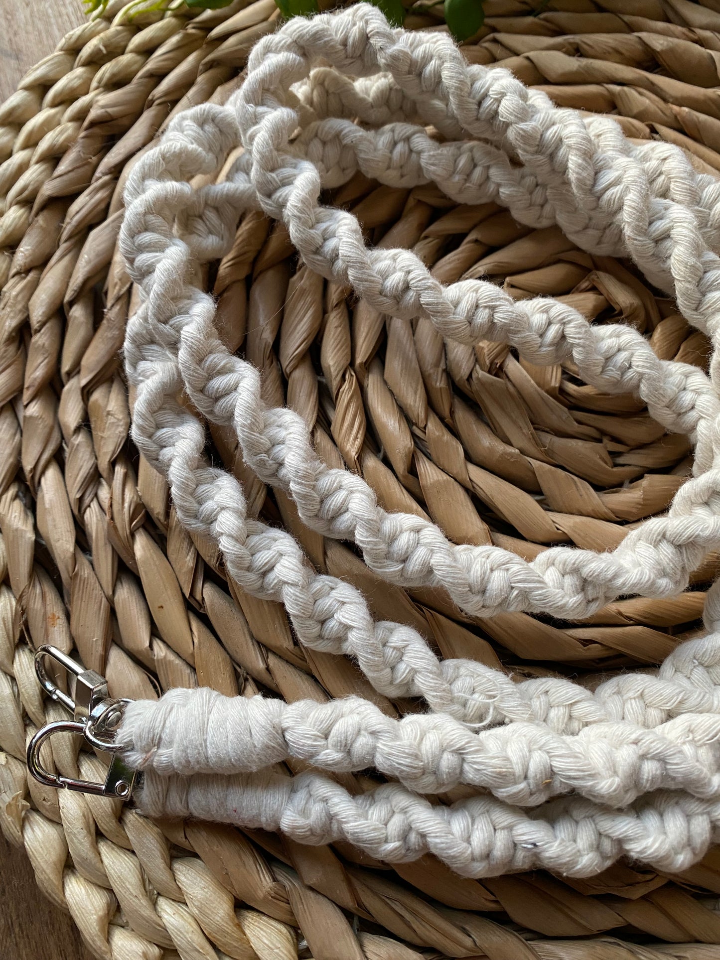 Close up of a macrame crossbody phone strap made in a twisted knot style, with natural yarn and silver clips on each end, on a wicker mat.