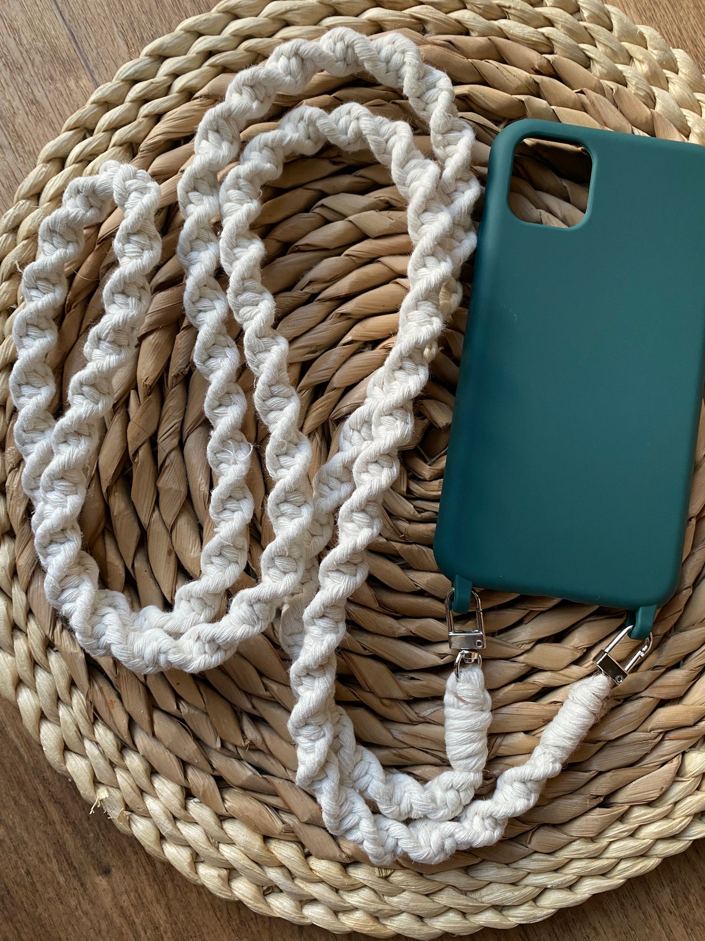 An empty green phone case sits on a woven wicker mat, with a long crossbody macrame strap attached to it with two silver clips at the bottom.