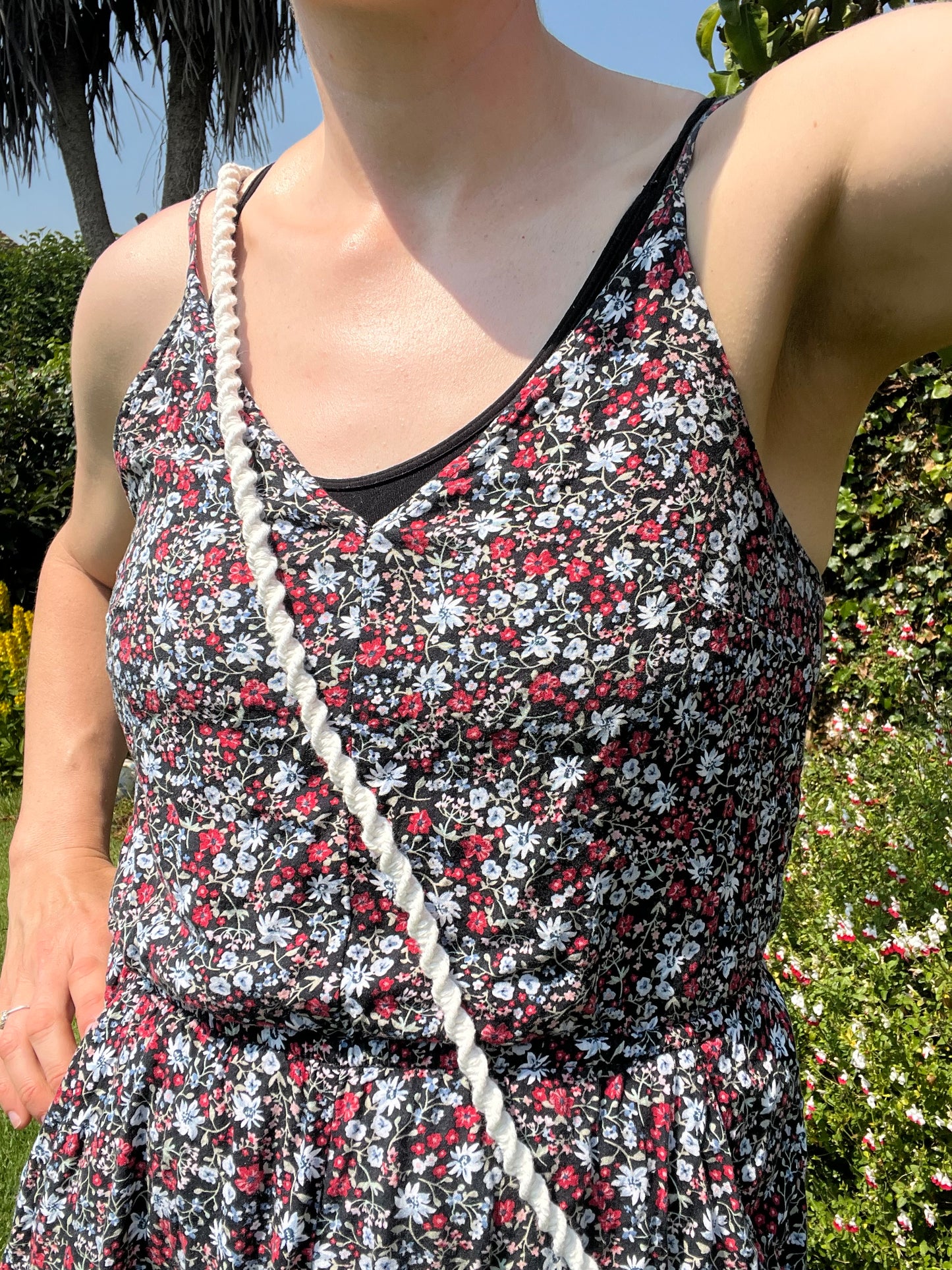 A person wearing a floral playsuit is standing outdoors in a garden under bright sunlight carrying a crossbody phone holder made with natural yarn across their chest.