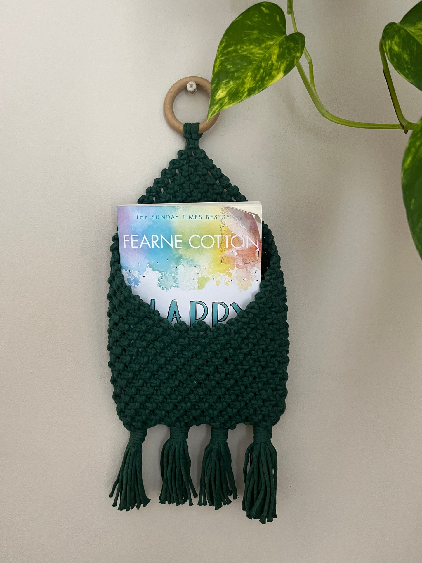 A green, handmade Macra-Made-With-Love Bedside storage pouch book sleeve with fringe hangs on a white wall by a wooden ring. Inside the pocket, a book titled "Happy" by Fearne Cotton is visible. To the right, a green plant adds natural decor. This recycled cotton decor piece is perfect for eco-friendly bedroom storage.