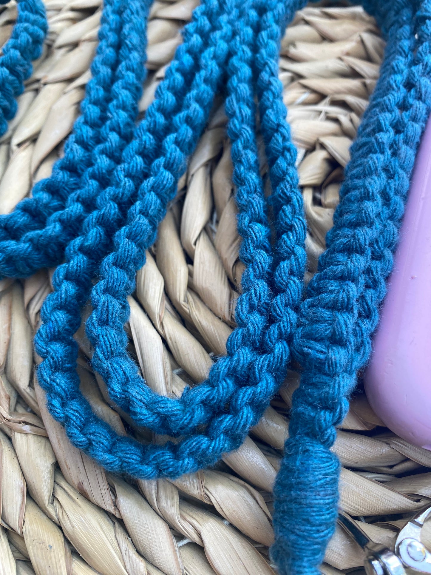 Close-up of a teal macramé cord intricately woven over a natural woven straw surface, showcasing an eco-friendly, handmade Recycled cotton crossbody phone strap by Macra-Made-With-Love. The edge of a light purple object is visible in the corner.