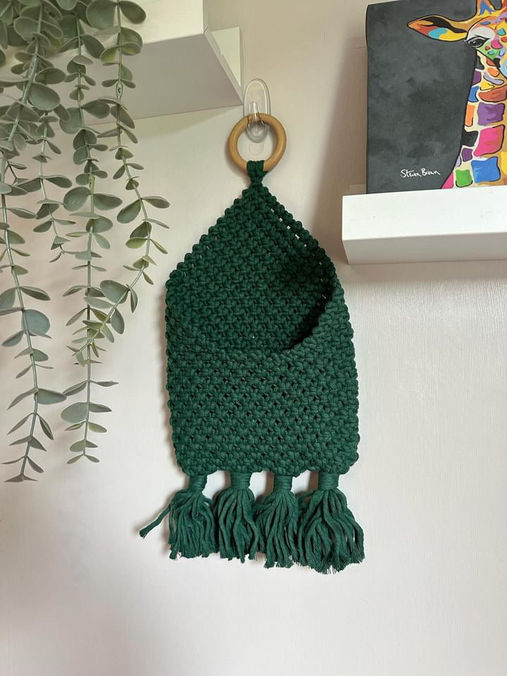 A dark green, handmade Macra-Made-With-Love Bedside Storage Pouch Book Sleeve with tassels at the bottom is suspended from a round wooden ring on a white wall. A vibrant, colorful giraffe painting sits on a shelf to the right, and a leafy plant hangs from the top left corner of the image.
