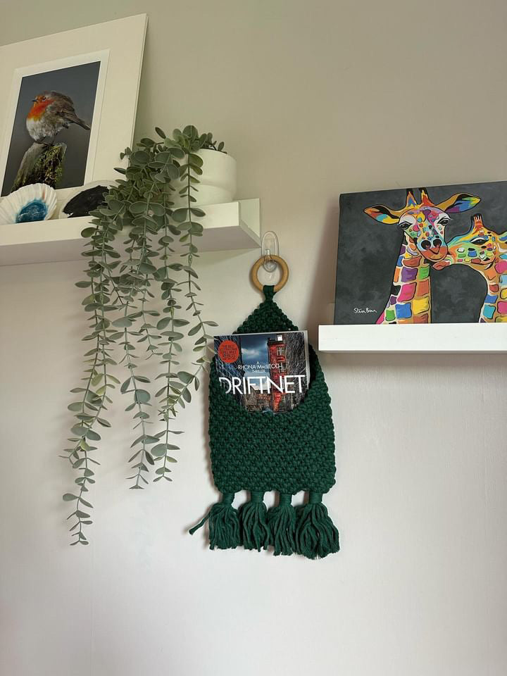 A green crocheted Bedside storage pouch book sleeve decor by Macra-Made-With-Love holds a book titled "Driftnet". It is hung on a white hook on a beige wall. Surrounding shelves display a potted plant, a framed bird photo, and a colorful painting of a giraffe.