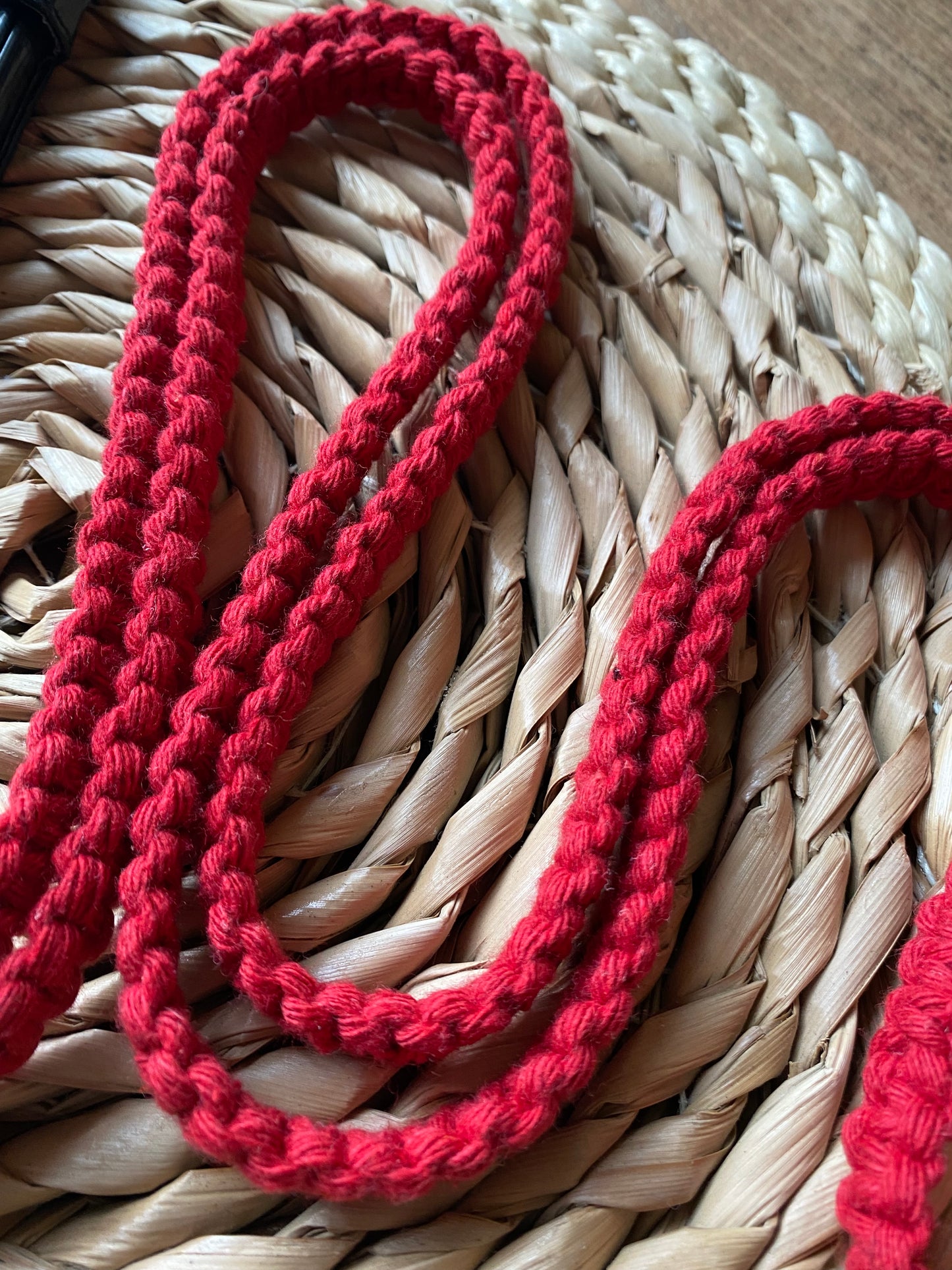 A close-up image of a red, braided cord neatly arranged on a woven, straw-like surface. This Recycled cotton crossbody phone strap by Macra-Made-With-Love displays a consistent pattern and contrasts beautifully against the natural texture of the background, making it an ideal eco-friendly phone accessory.