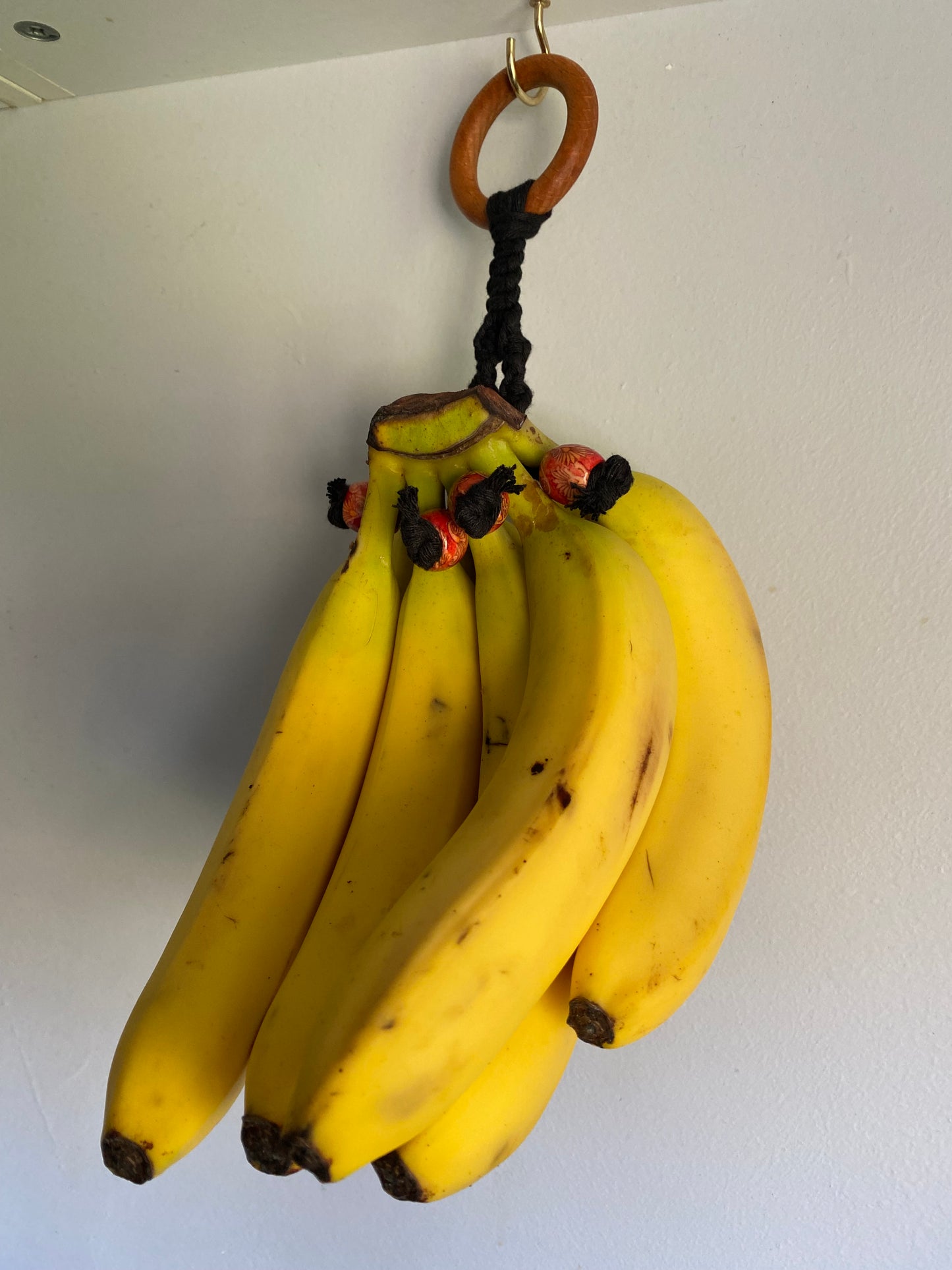 A bunch of ripe yellow bananas hangs on a handmade wooden banana holder against a white wall. The bananas, with a few dark spots indicating ripeness, are kept suspended by a black cord attached to the Kitchen Space Saving Eco Friendly Banana Hanger from Macra-Made-With-Love.