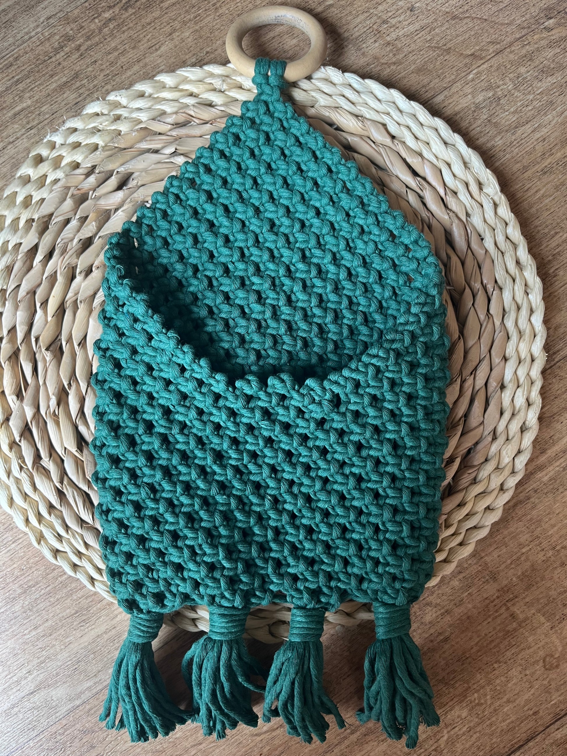A green macrame wall hanging featuring a cylindrical wooden ring at the top. The handmade **Bedside storage pouch book sleeve** by **Macra-Made-With-Love** showcases a diamond-shaped pocket and four fringes at the bottom, beautifully displayed on a round, woven placemat with alternating light and dark straw colors.