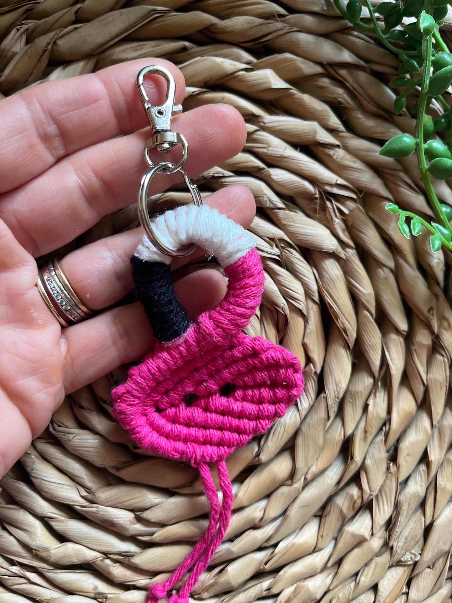 Handmade macrame keychain in the shape of a flamingo, made using fuchsia pink recycled cotton, on a metal split ring with lobster clasp. The keyring is displayed on a round wicker surface next to some green plant vines.