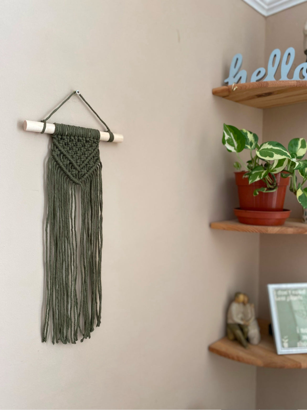 Handmade macrame wall hanging, made with green recycled cotton, on a solid wooden dowel. The wall hanging is displayed hanging on a plain painted wall, with corner shelves visible to the right.