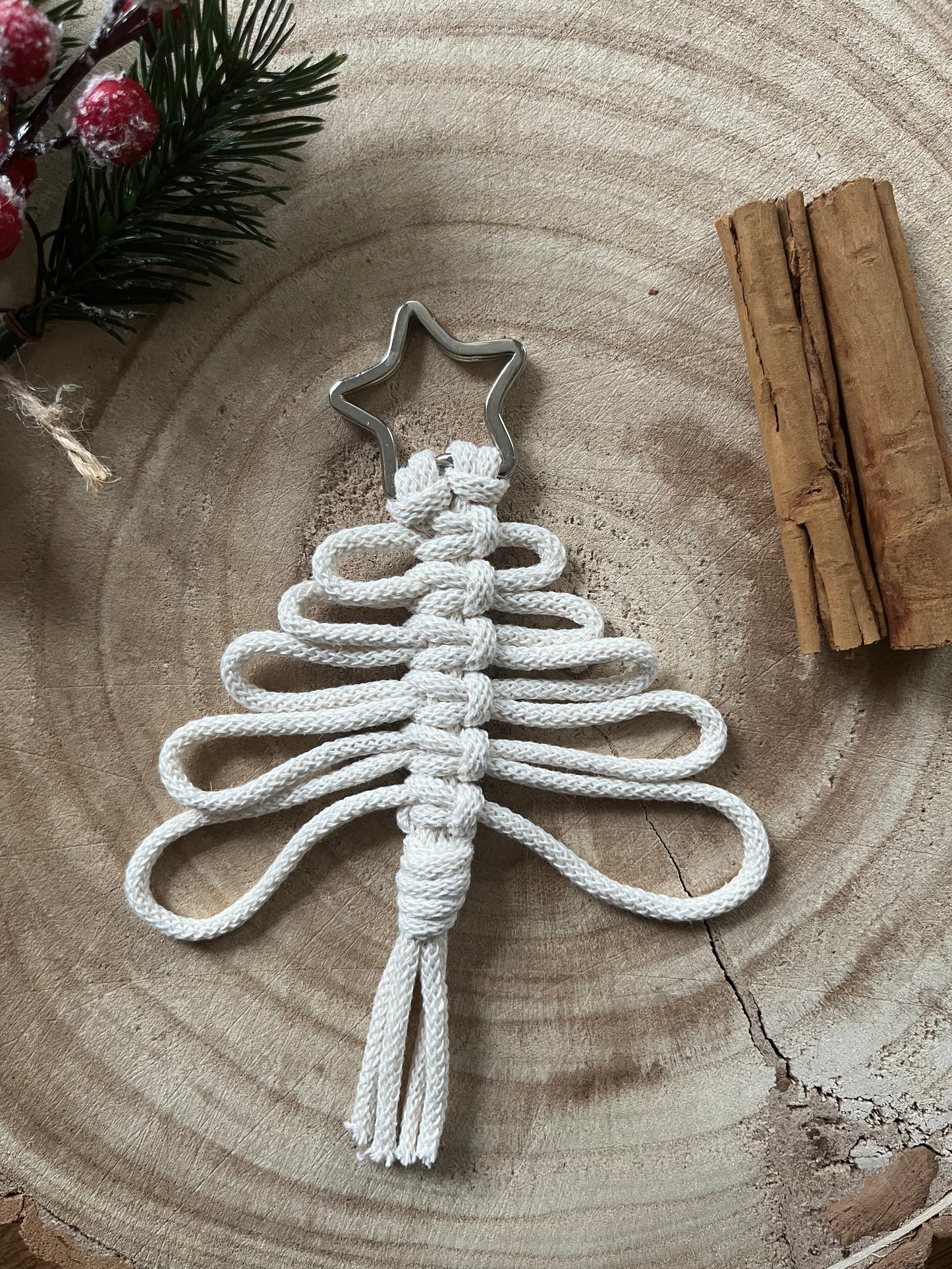 The Knotted Yarn Hanging Christmas Tree Decorations by Macra-Made-With-Love are beautifully handcrafted from white rope, shaped into a tree with looping branches. This ornament, set on a wooden surface and topped with a metal star, is accompanied by two cinnamon sticks and a sprig of evergreen with red berries—making it an ideal piece of handmade holiday decor.