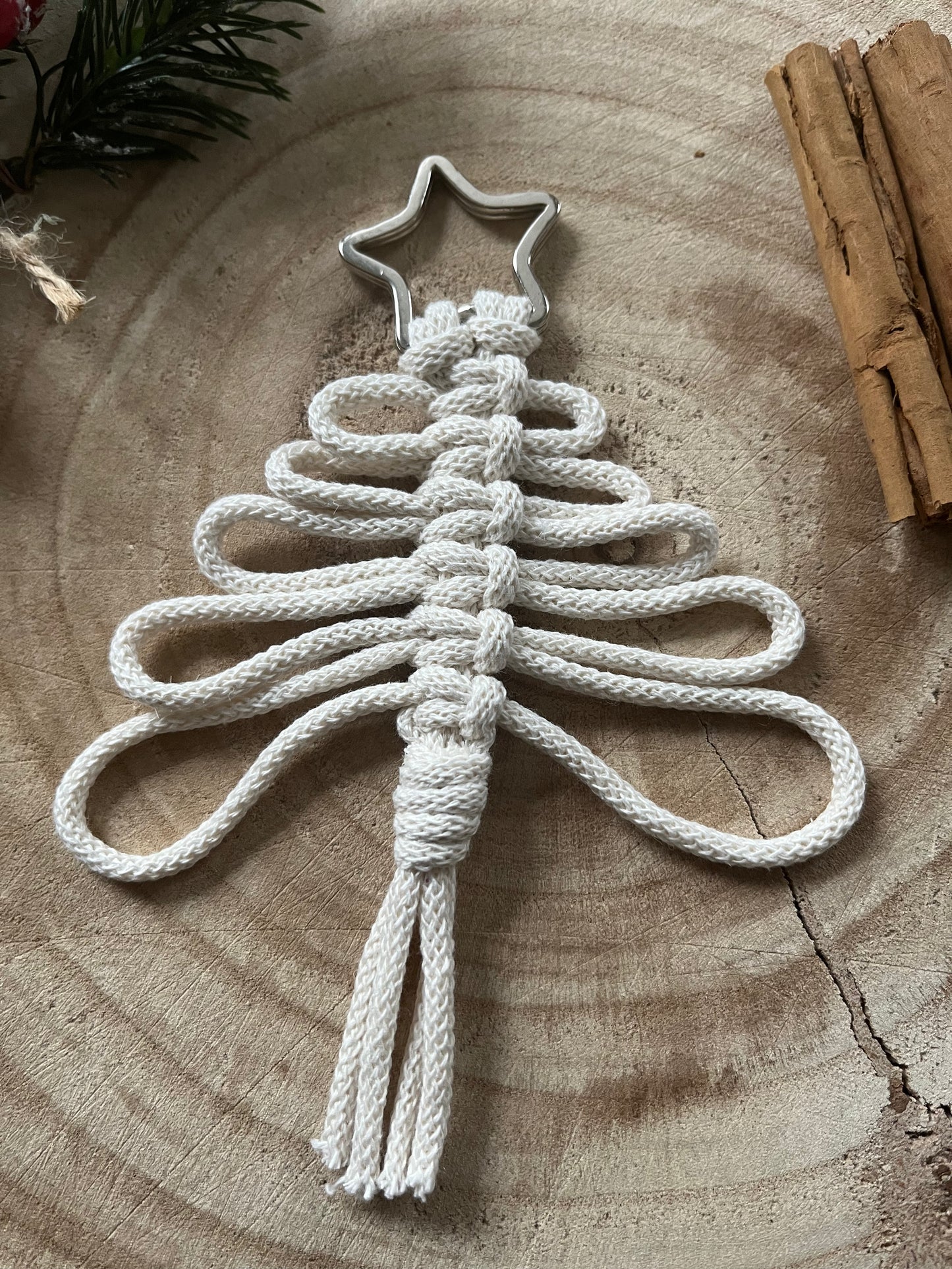A Knotted Yarn Hanging Christmas Tree Decoration from Macra-Made-With-Love, crafted with white cord and embellished with a silver star at the top, rests on a wooden surface. The ornament showcases a looping, woven design and ends in a tassel at the bottom. Adding to the festive atmosphere are nearby sticks of cinnamon and sprigs of greenery, enhancing this handmade holiday décor item.
