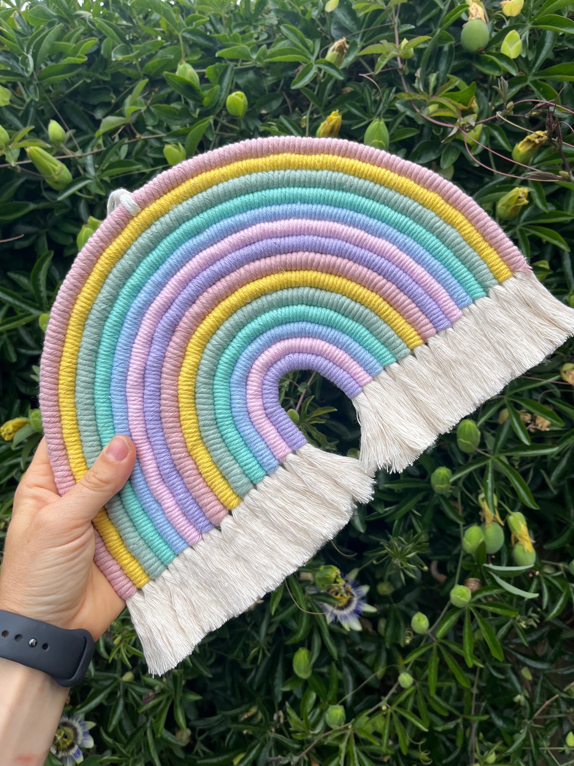 A hand holding an XL macrame rainbow outside, made with pastel rainbow shades of recycled cotton yarn.