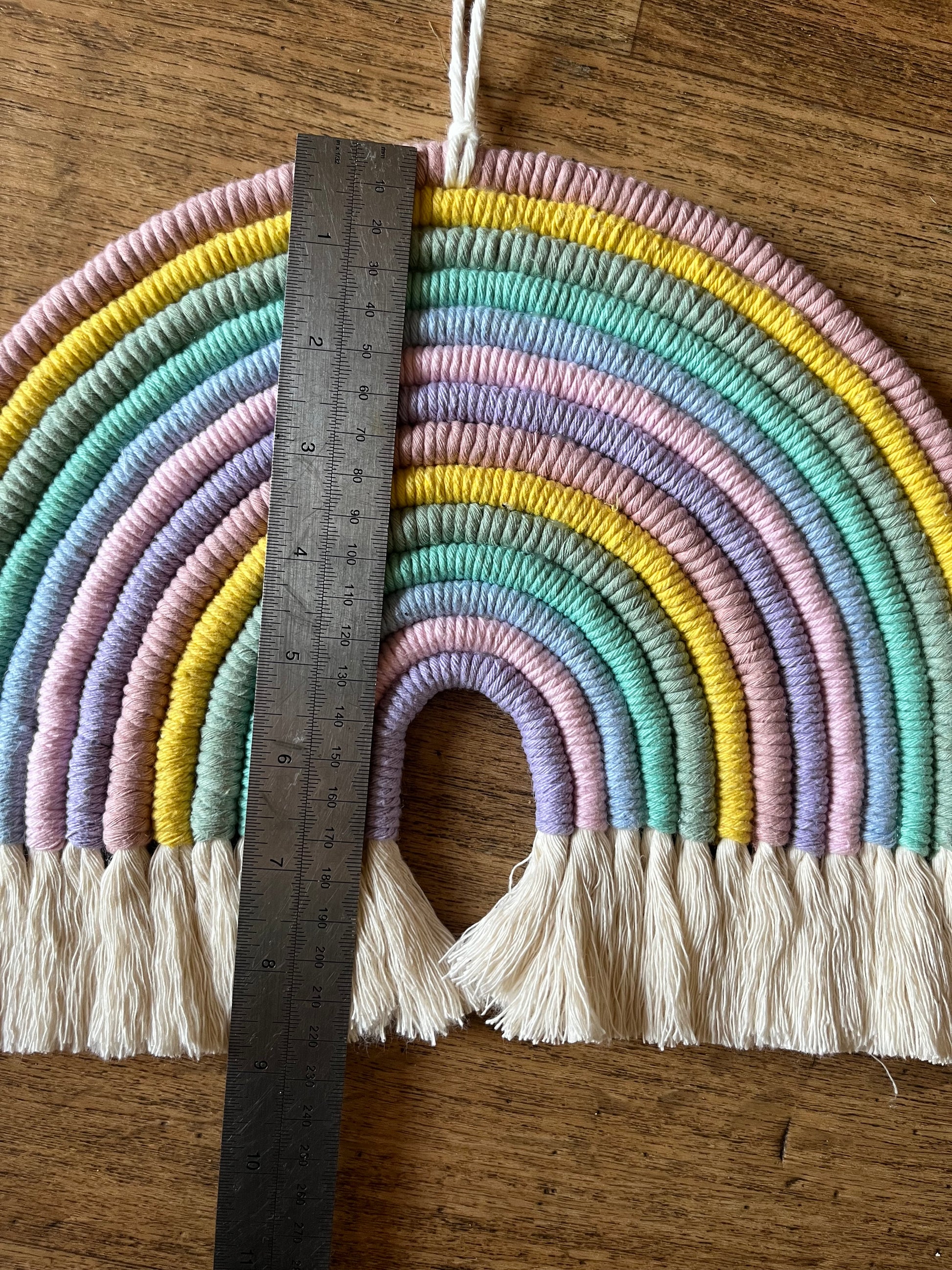 Handmade macrame rainbow wall hanging, made with recycled cotton. Shown laid out with a metal rule giving dimensions.