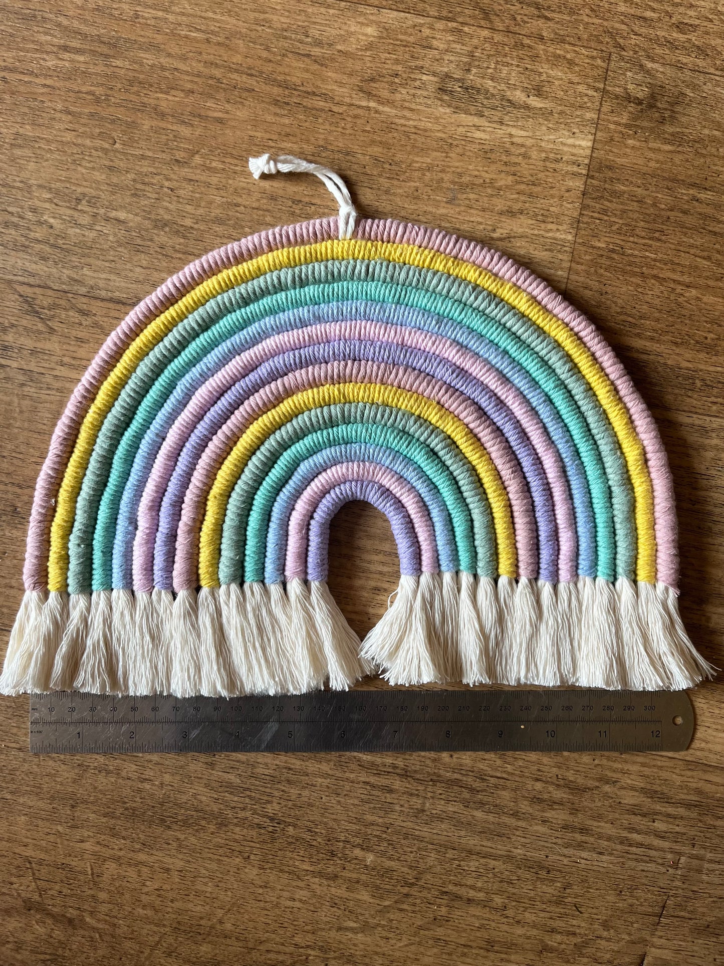 Handmade macrame rainbow wall hanging, shown laid on a wooden surface next to a metal rule.
