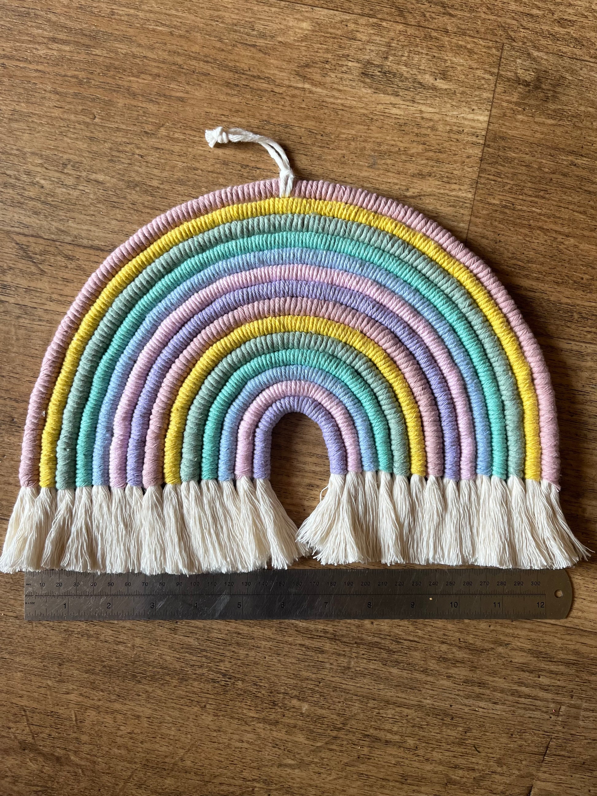 Handmade macrame rainbow wall hanging, shown laid on a wooden surface next to a metal rule.