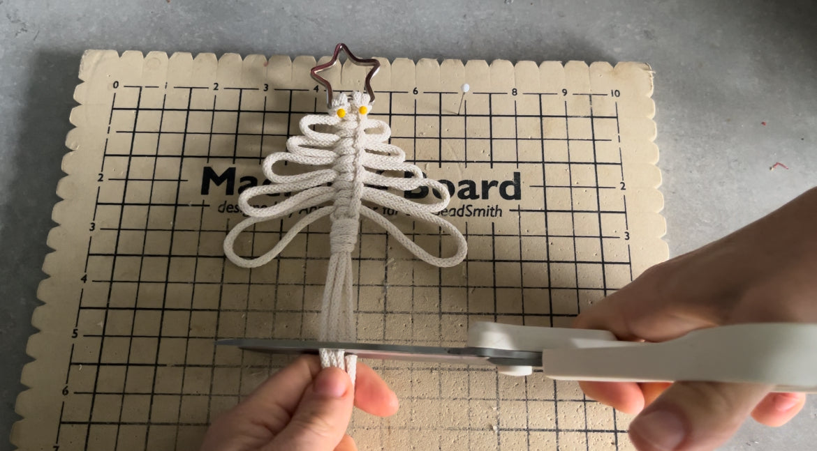 Hands using white scissors to cut the tassels of a handmade macramé Christmas tree. The tree, crafted with intricate macramé knots and adorned with a star at the top, is pinned to a crafting board.