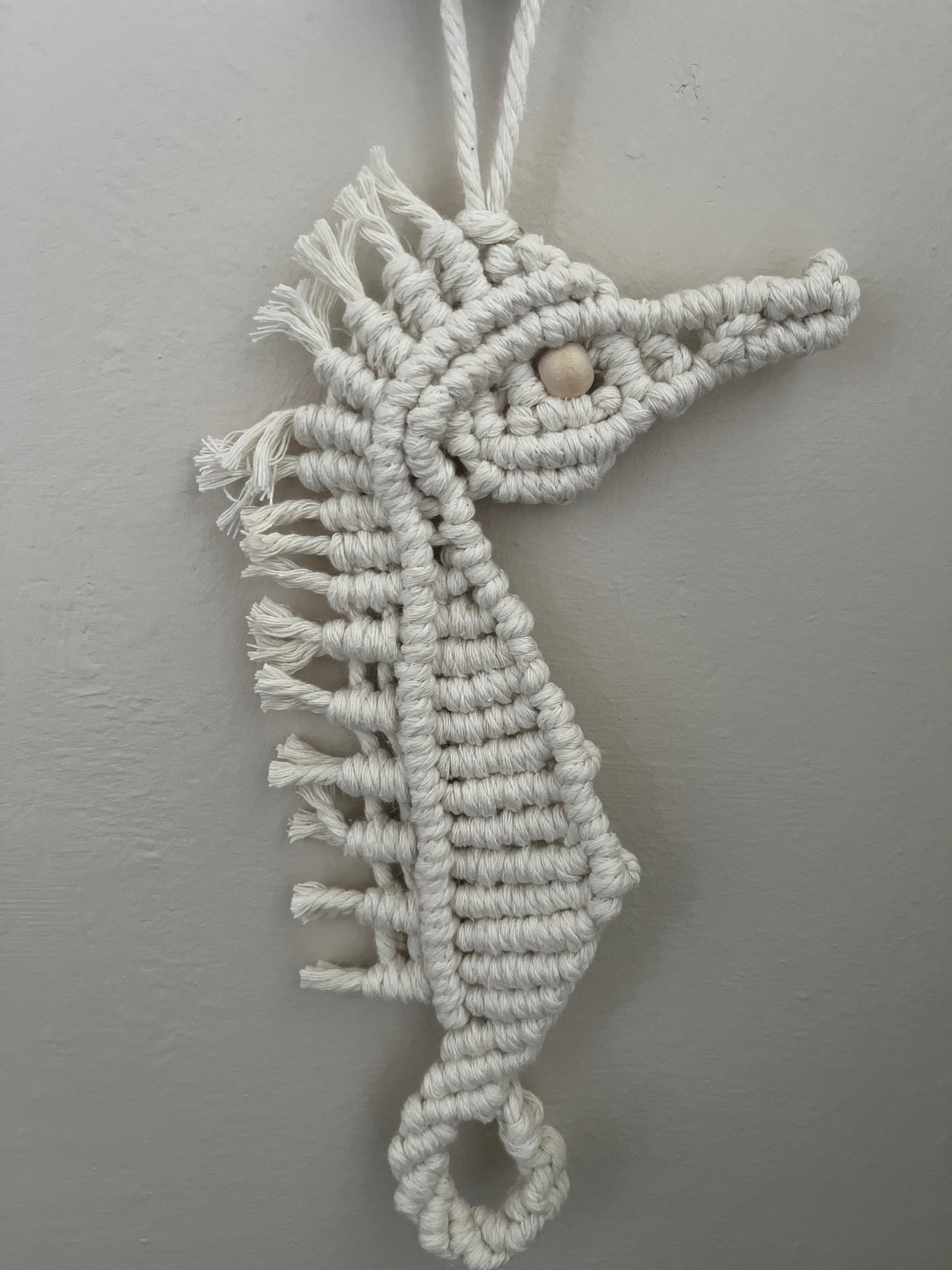 Macrame Seahorse shaped wall hanging decoration, handmade using natural recycled cotton yarn, and a small Woden bead, displayed hanging against a plain painted wall