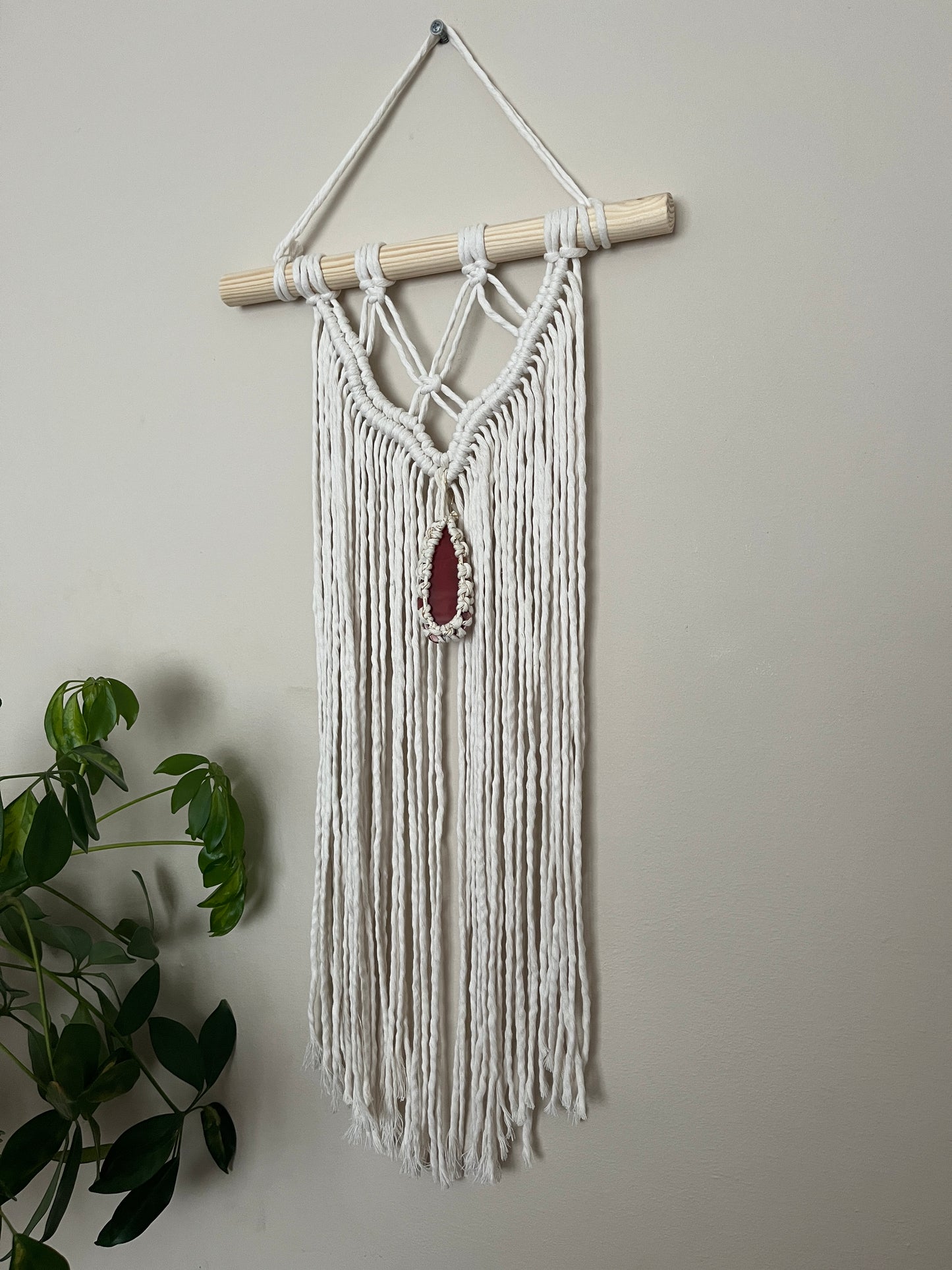 Macrame Wall Hanging, made with natural recycled cotton yarn on a wooden dowel, featuring a red agate slice. Displayed hanging against a plain painted wall, with a green plant visible in the bottom left corner.