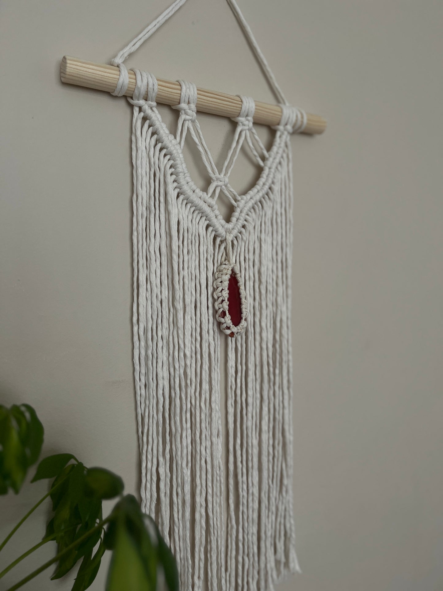 Macrame Wall Hanging, made with natural recycled cotton yarn on a wooden dowel, featuring a red agate slice. Displayed hanging against a plain painted wall, with a green plant visible in the bottom left corner.