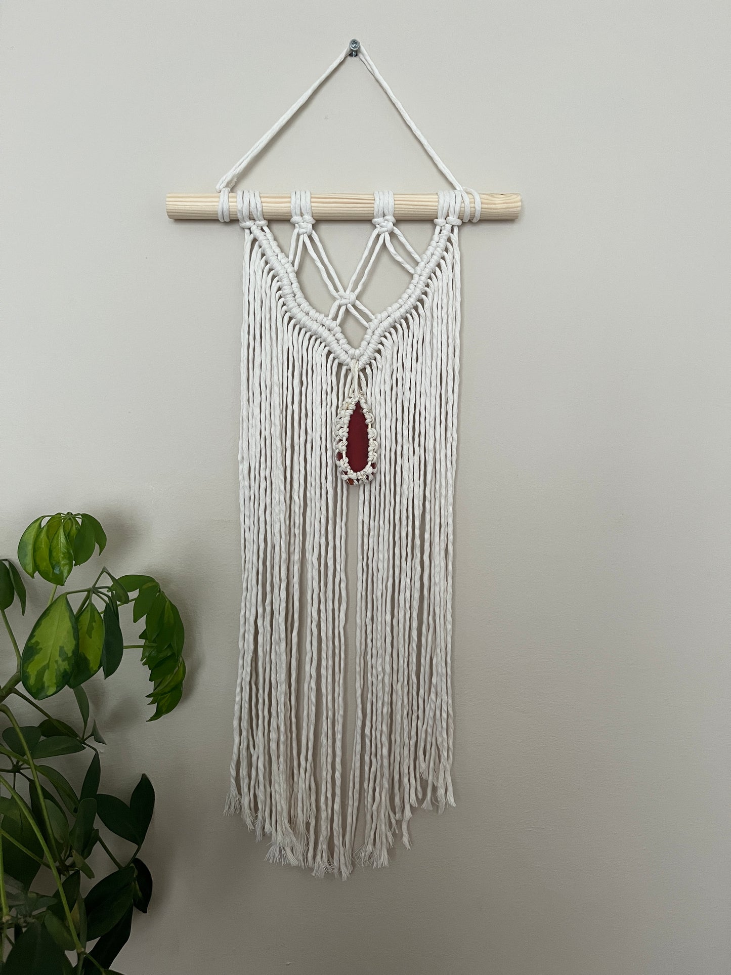 Macrame Wall Hanging, made with natural recycled cotton yarn on a wooden dowel, featuring a red agate slice. Displayed hanging against a plain painted wall, with a green plant visible in the bottom left corner.