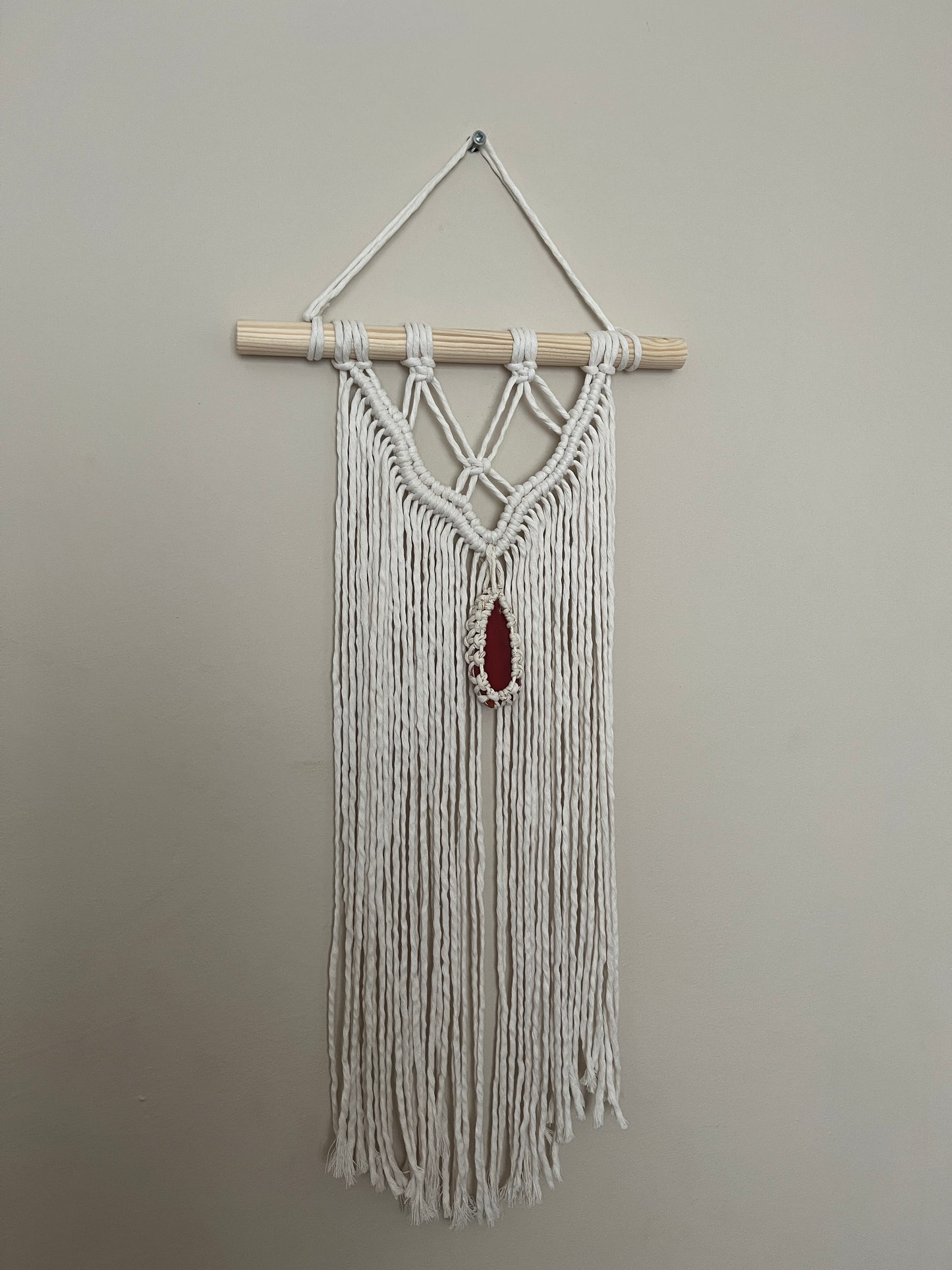 Macrame Wall Hanging, made with natural recycled cotton yarn on a wooden dowel, featuring a red agate slice. Displayed hanging against a plain painted wall.