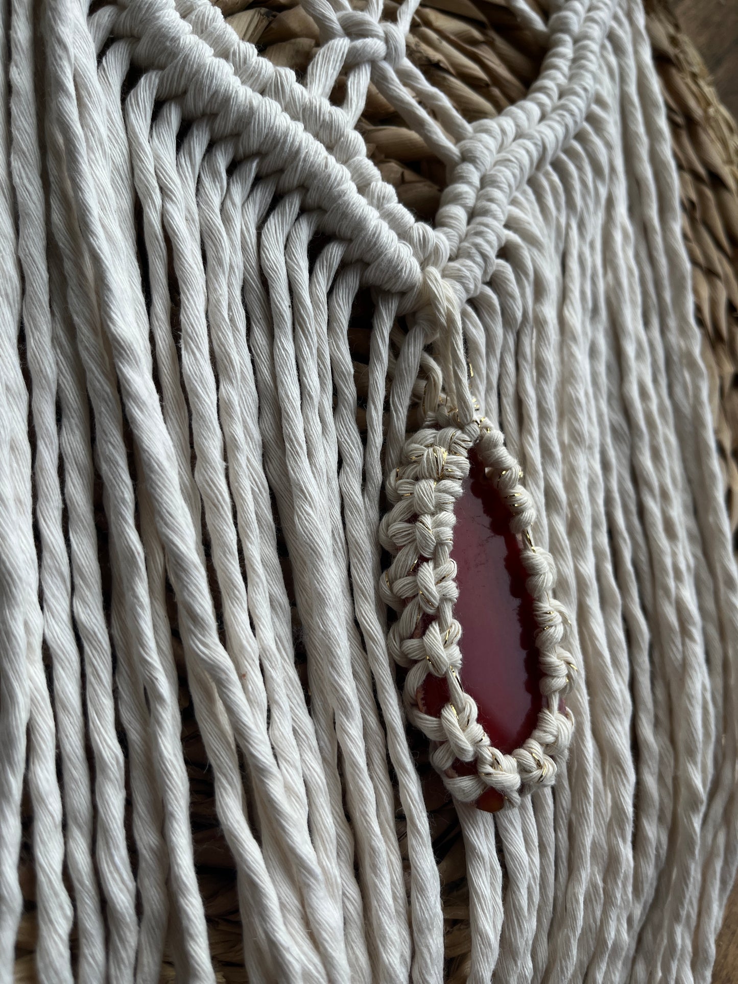 Close up of a Macrame Wall Hanging, made with natural recycled cotton yarn on a wooden dowel, featuring a red agate slice. Displayed laid flat on a wicker mat.