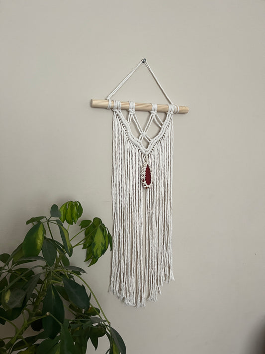Macrame Wall Hanging, made with natural recycled cotton yarn on a wooden dowel, featuring a red agate slice. Displayed hanging against a plain painted wall, with a green plant visible in the bottom left corner.