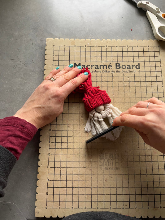 A person's hands are shown crafting a macrame Santa decoration on a macrame board, with cotton cords and wooden bead.